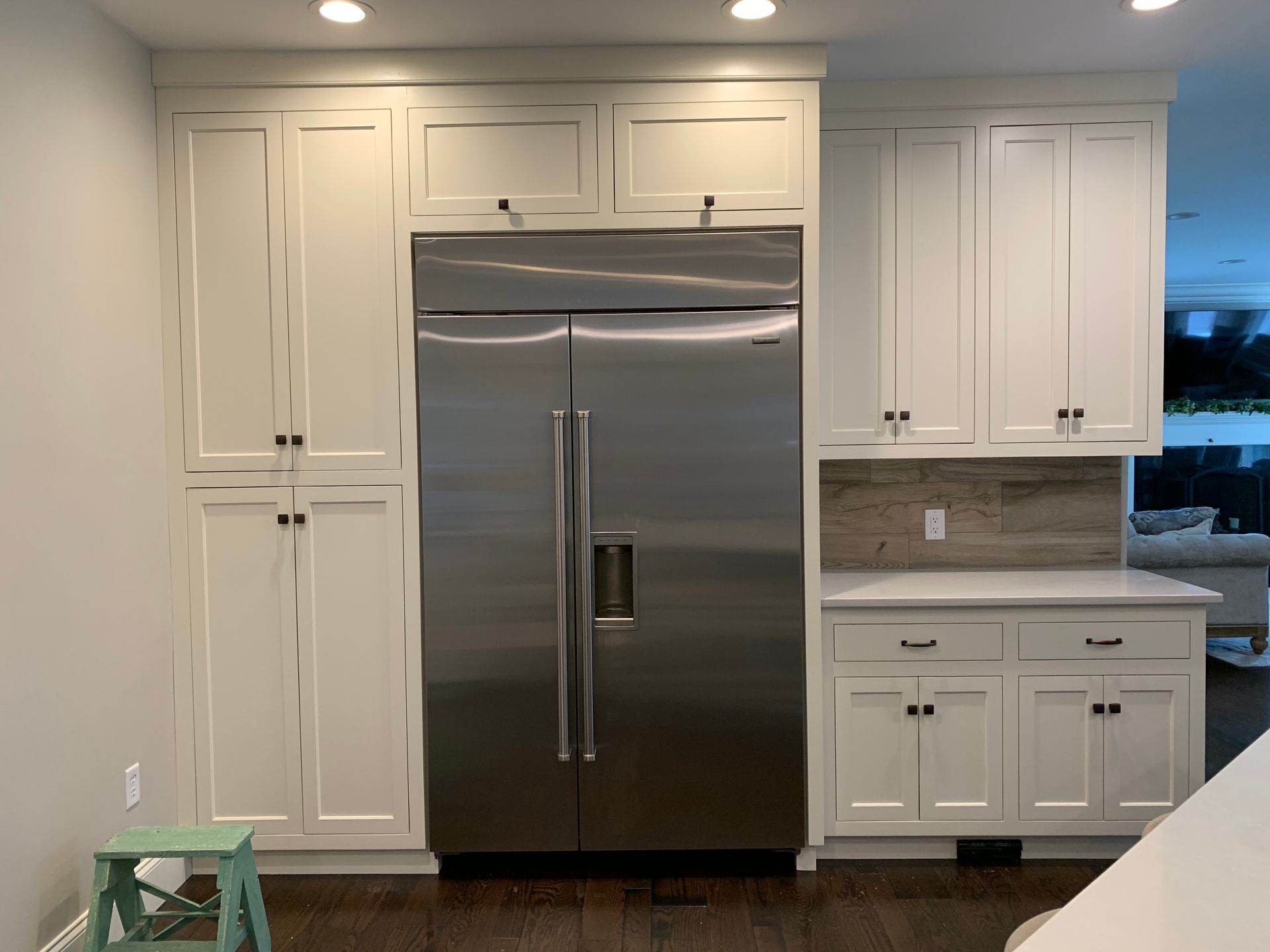 A stainless steel refrigerator sits between white cabinet units in a kitchen with hardwood floors and a tiled backsplash.
