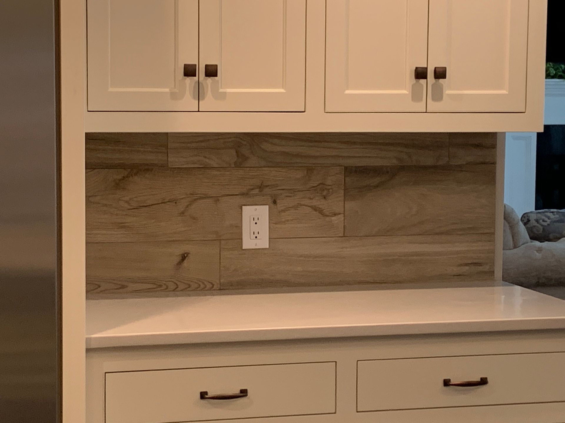 A kitchen nook with white cabinets, a light wood-look tiled backsplash, a white countertop, and a centered wall outlet.