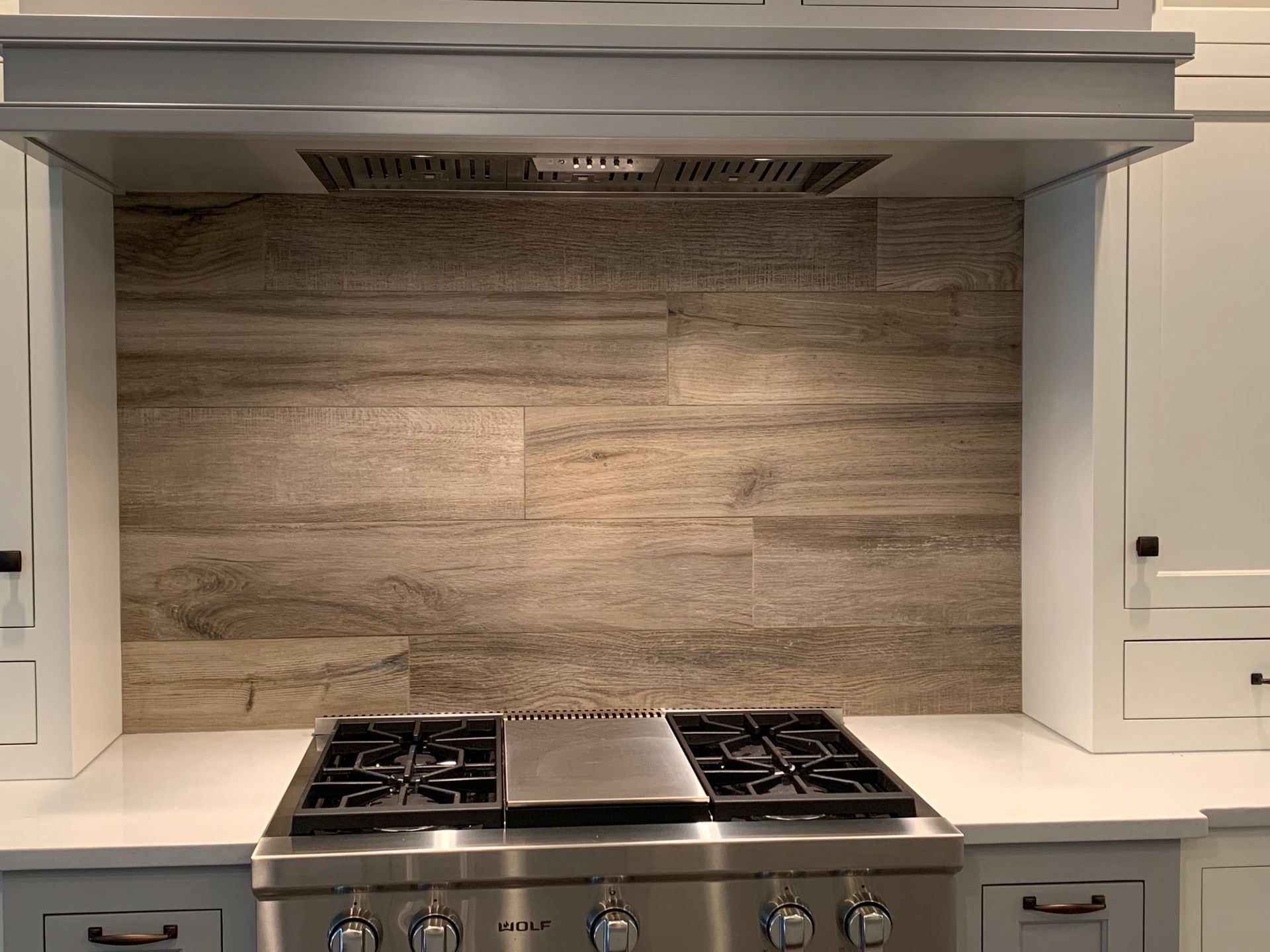A stainless steel gas range with a center griddle, set against a wood-look tiled backsplash under a grey range hood.