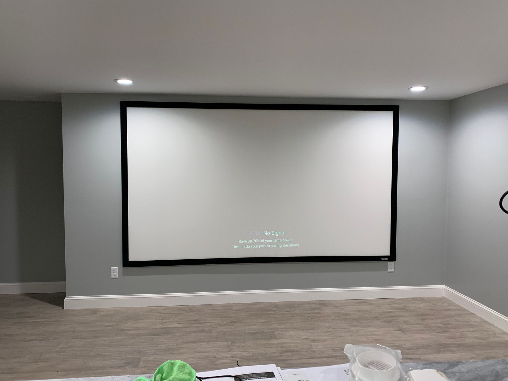 A large rectangular projector screen with a black frame mounted on a light gray wall in a room with wood-look flooring.