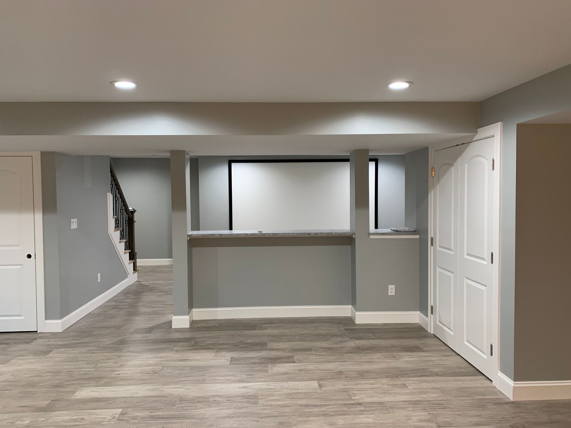 Finished basement with light gray walls, wood-look flooring, a white pass-through counter, and stairs to the upper level.