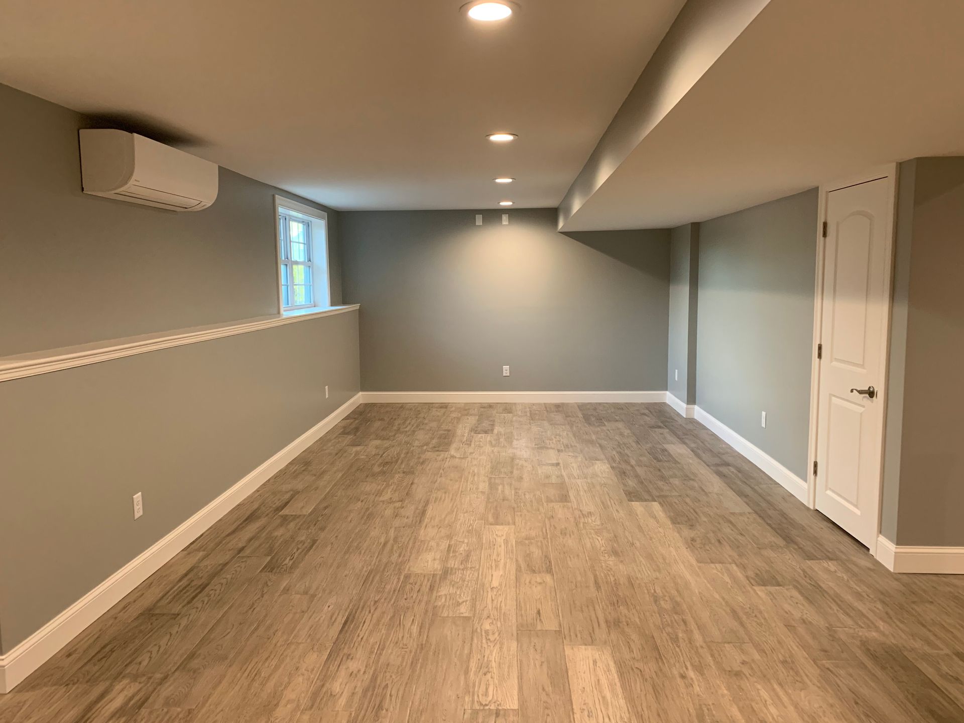 A renovated, empty basement with grey walls, light wood-look flooring, recessed lighting, and a white interior door.