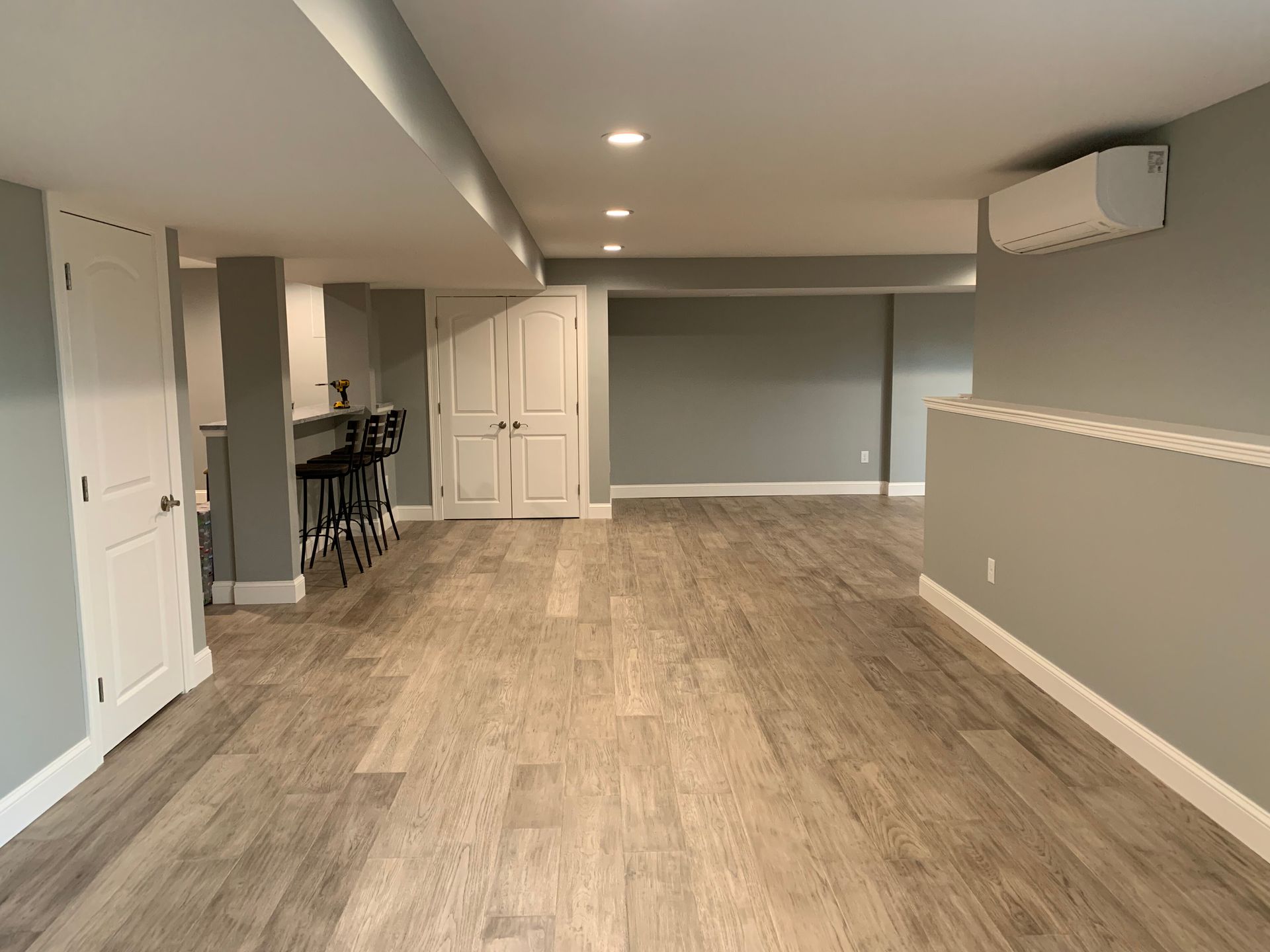 A finished basement with light gray walls, wood-look flooring, recessed lighting, a small bar area, and a wall AC unit.