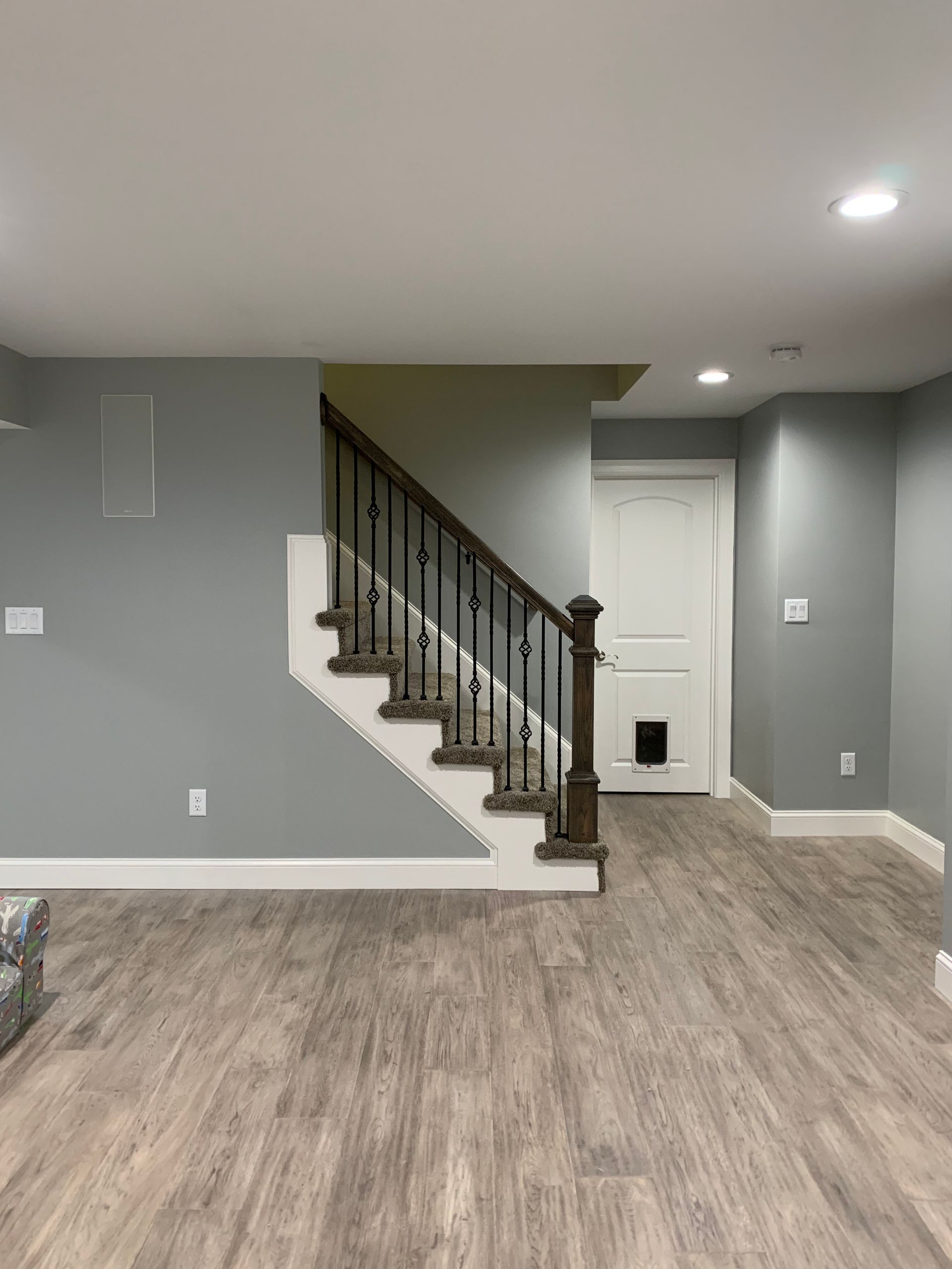 A modern basement room featuring light gray walls, wood-look flooring, and a staircase with dark railings and white trim.