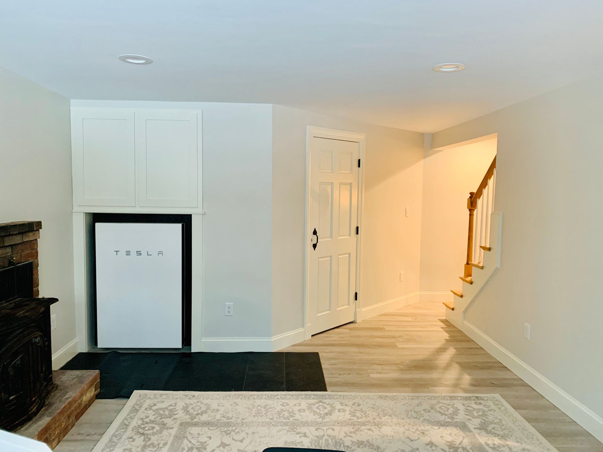 A basement room featuring a white Tesla Powerwall mounted in a wall alcove, next to a doorway and staircase with wood steps.