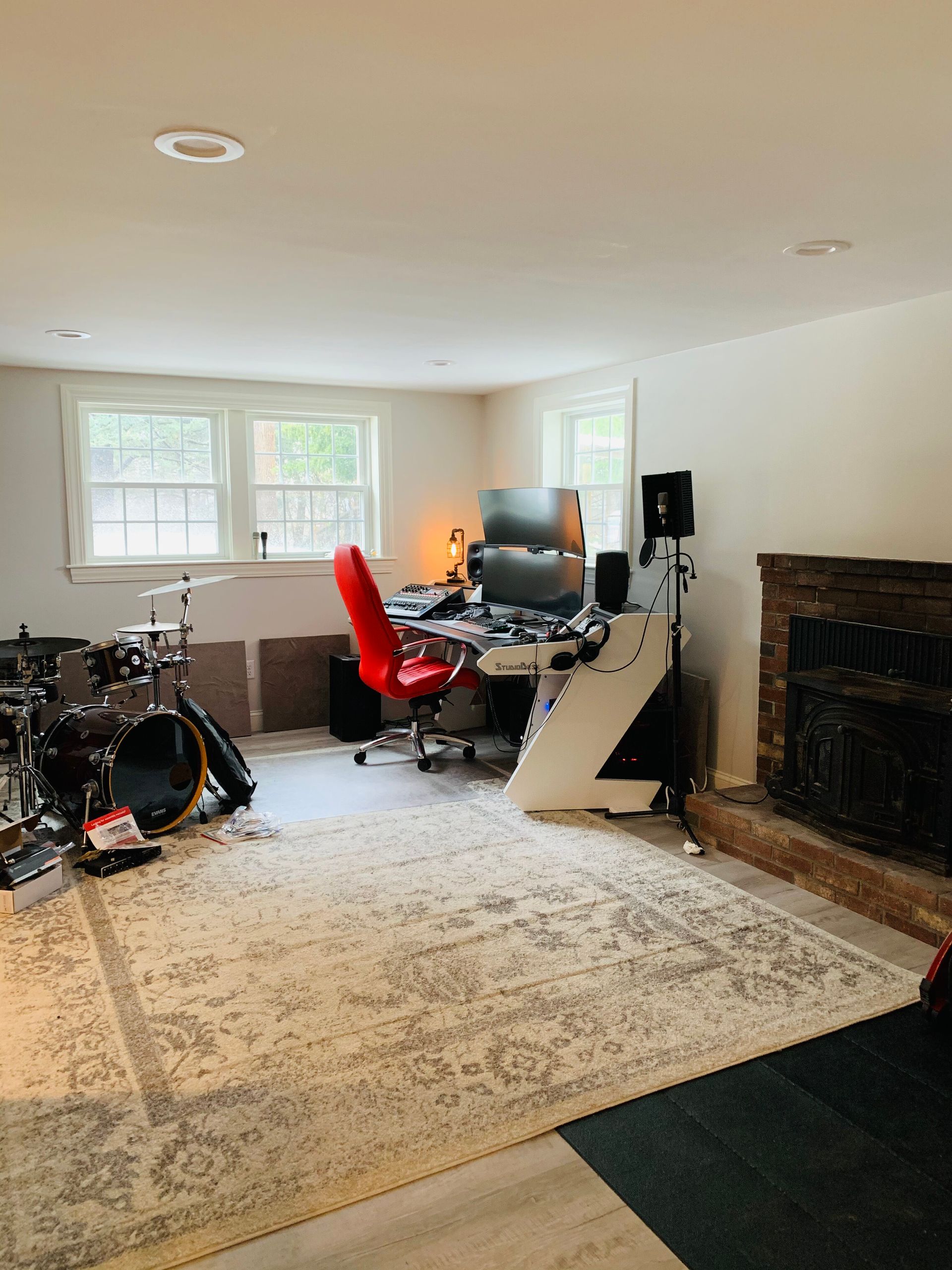 A home studio featuring a drum set on the left, a white computer desk with a red chair, and a brick fireplace on the right.