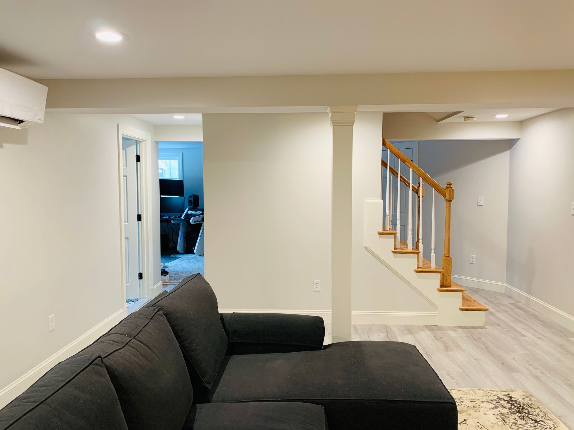 A finished basement room with a dark sectional sofa, neutral gray walls, light wood flooring, and stairs leading upward.