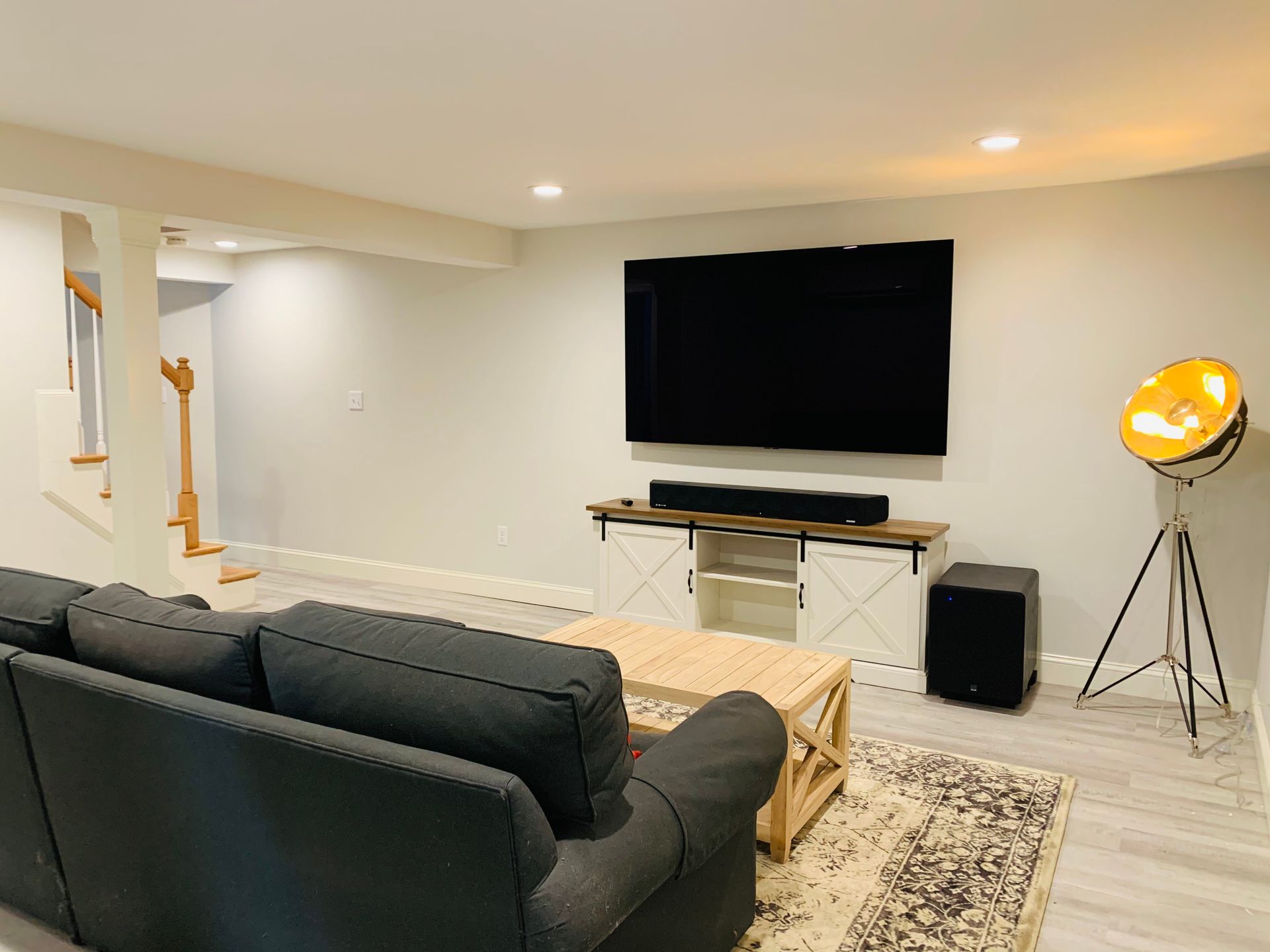 A modern living room with a large wall-mounted TV above a white media console, a grey sofa, and a tripod spotlight lamp.