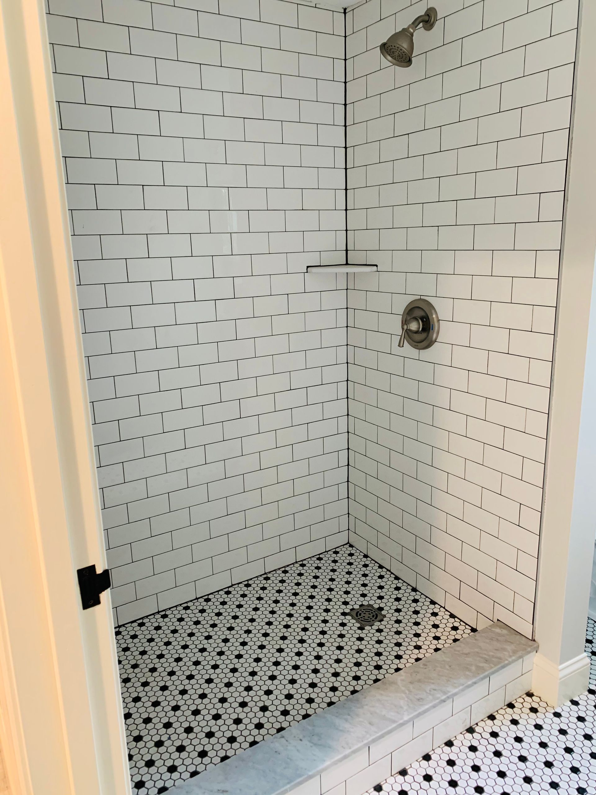 A walk-in shower with white brick-patterned wall tiles and a black-and-white patterned tile floor and threshold.
