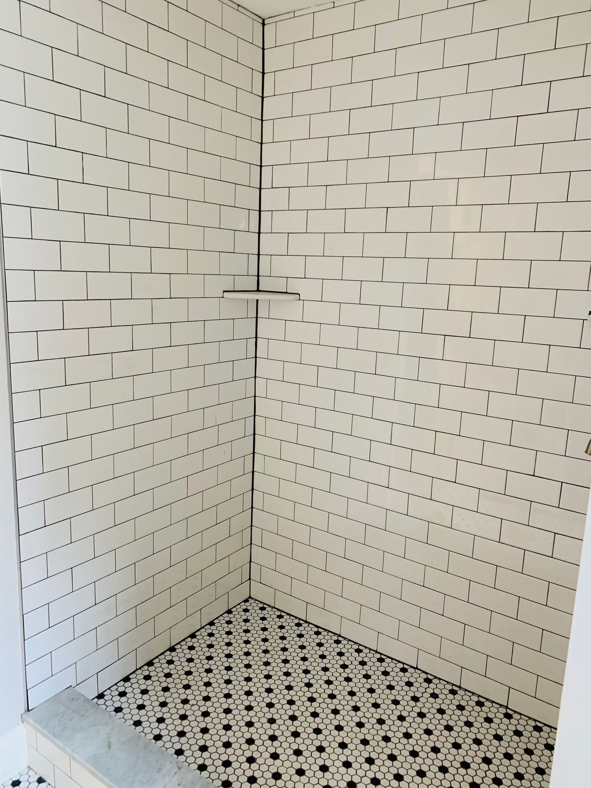 A corner shower with white subway tiles featuring dark grout and a patterned black-and-white tiled floor.