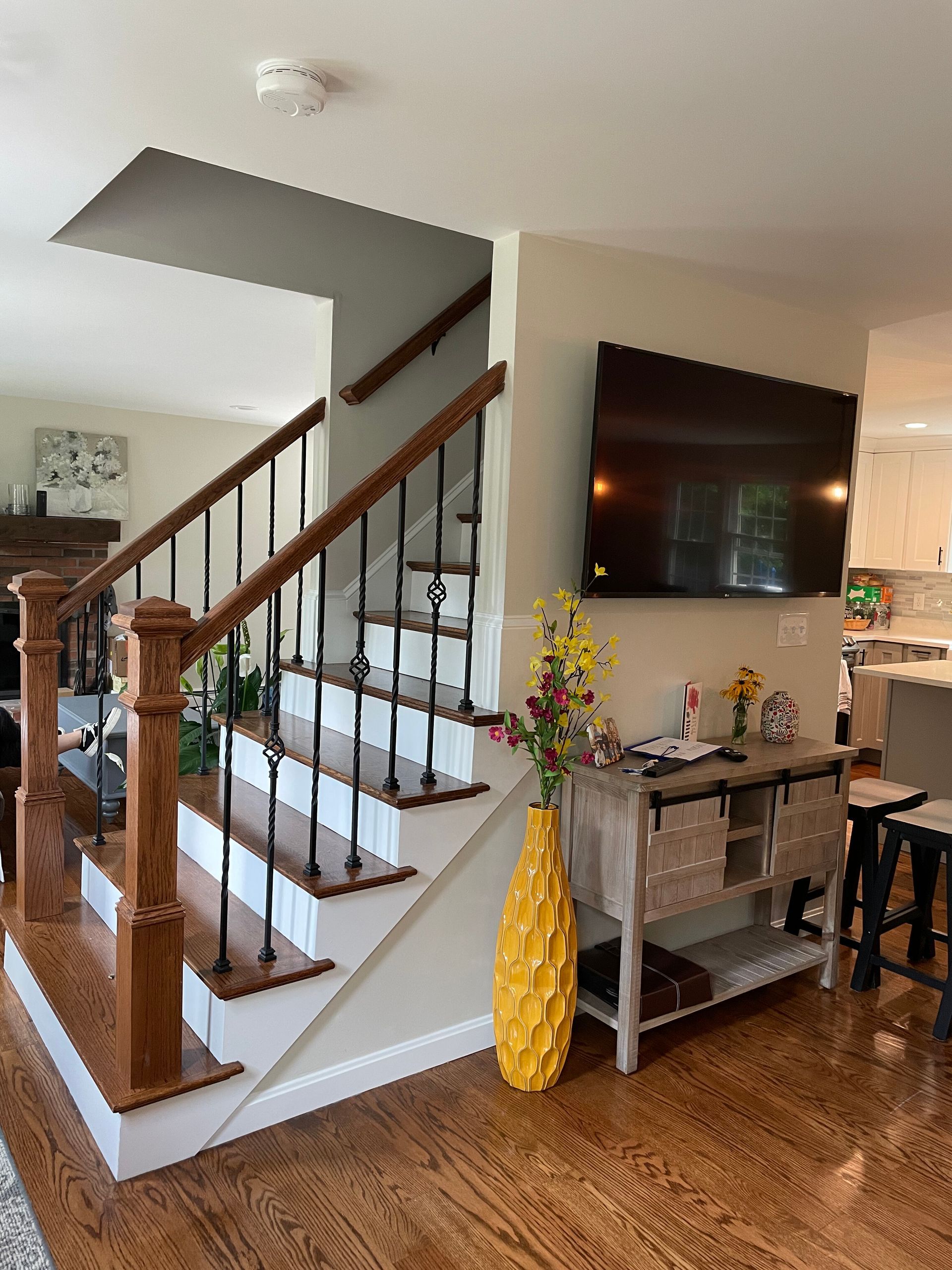 A wooden staircase with black iron railings next to a wall-mounted TV and a console table with a large yellow vase.