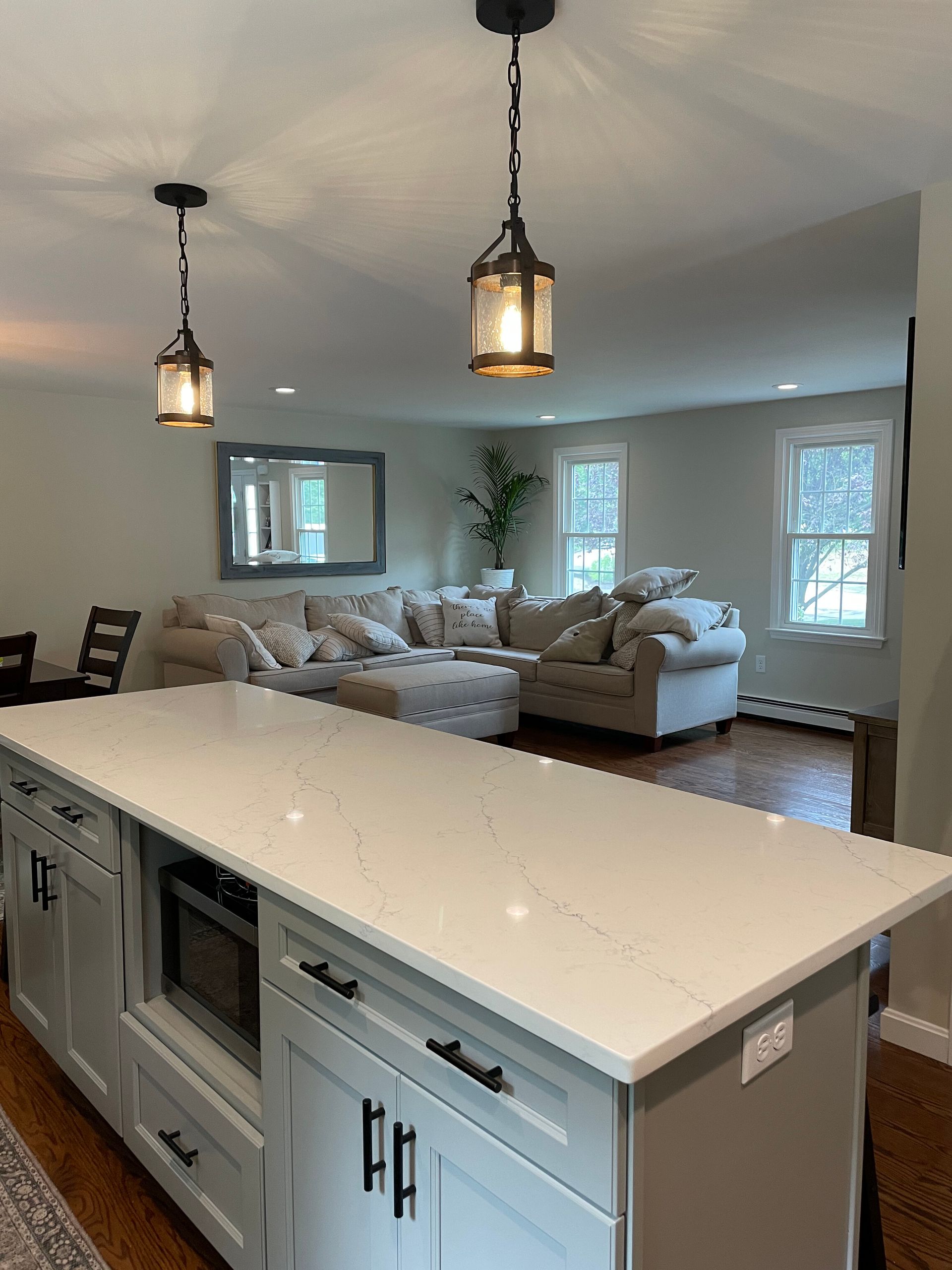 A kitchen island with a white countertop and grey cabinets foregrounds a living room with a sectional sofa and mirror.