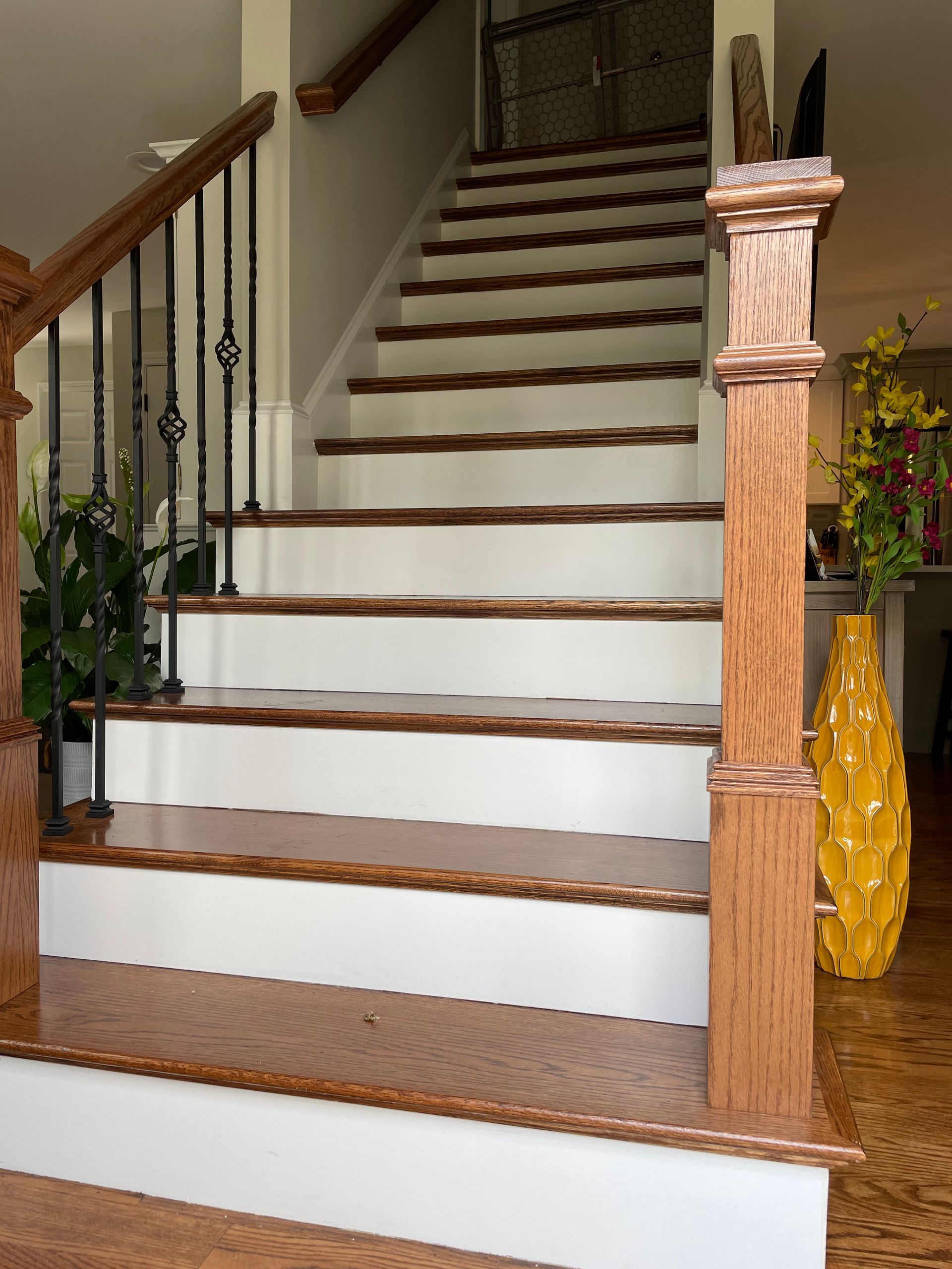 A staircase with white risers, wooden treads, and a wooden banister with black metal spindles in a brightly lit entryway.
