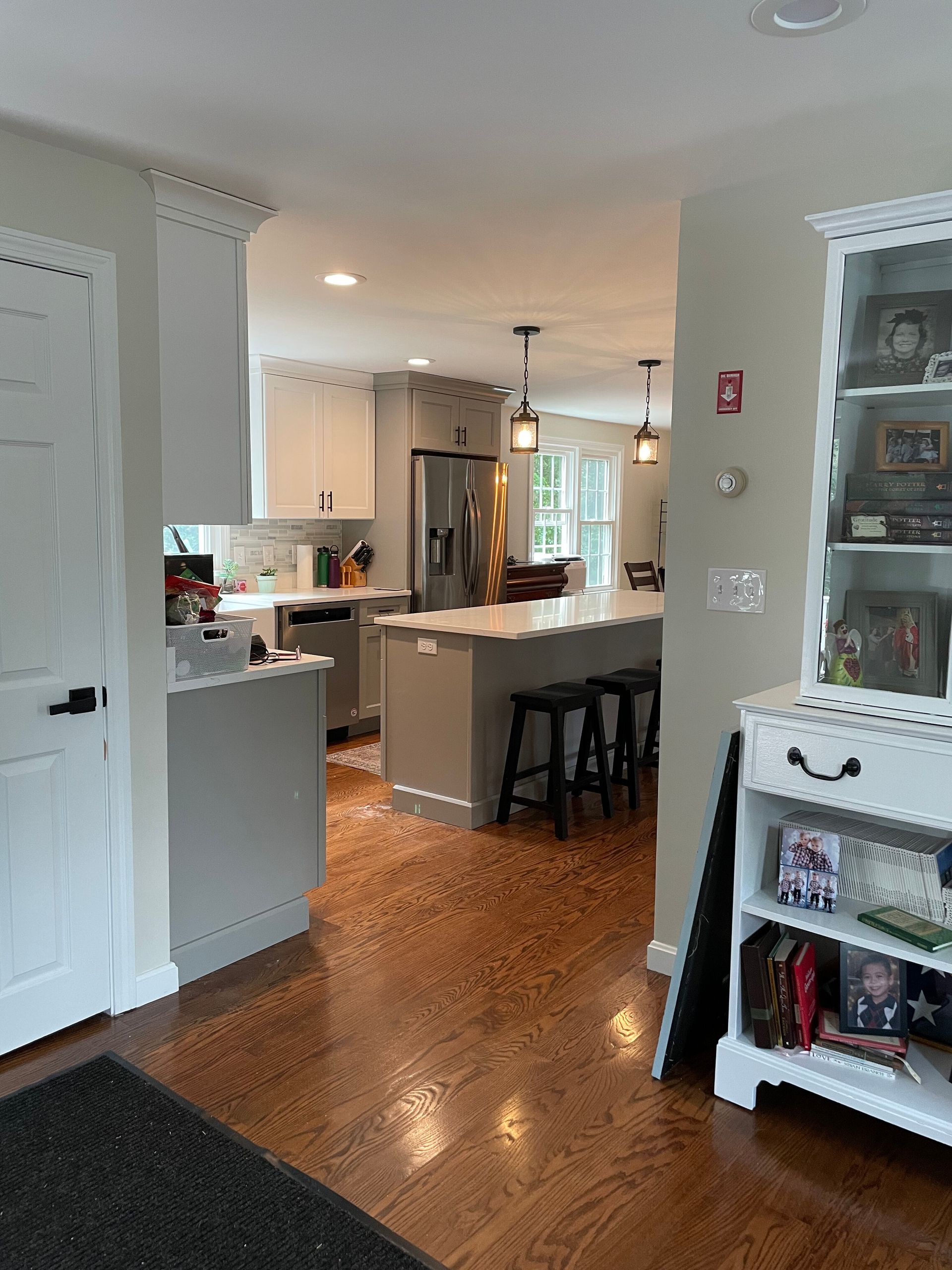 A view into a kitchen with grey cabinetry, a central island with black stools, and an open entryway with hardwood floors.
