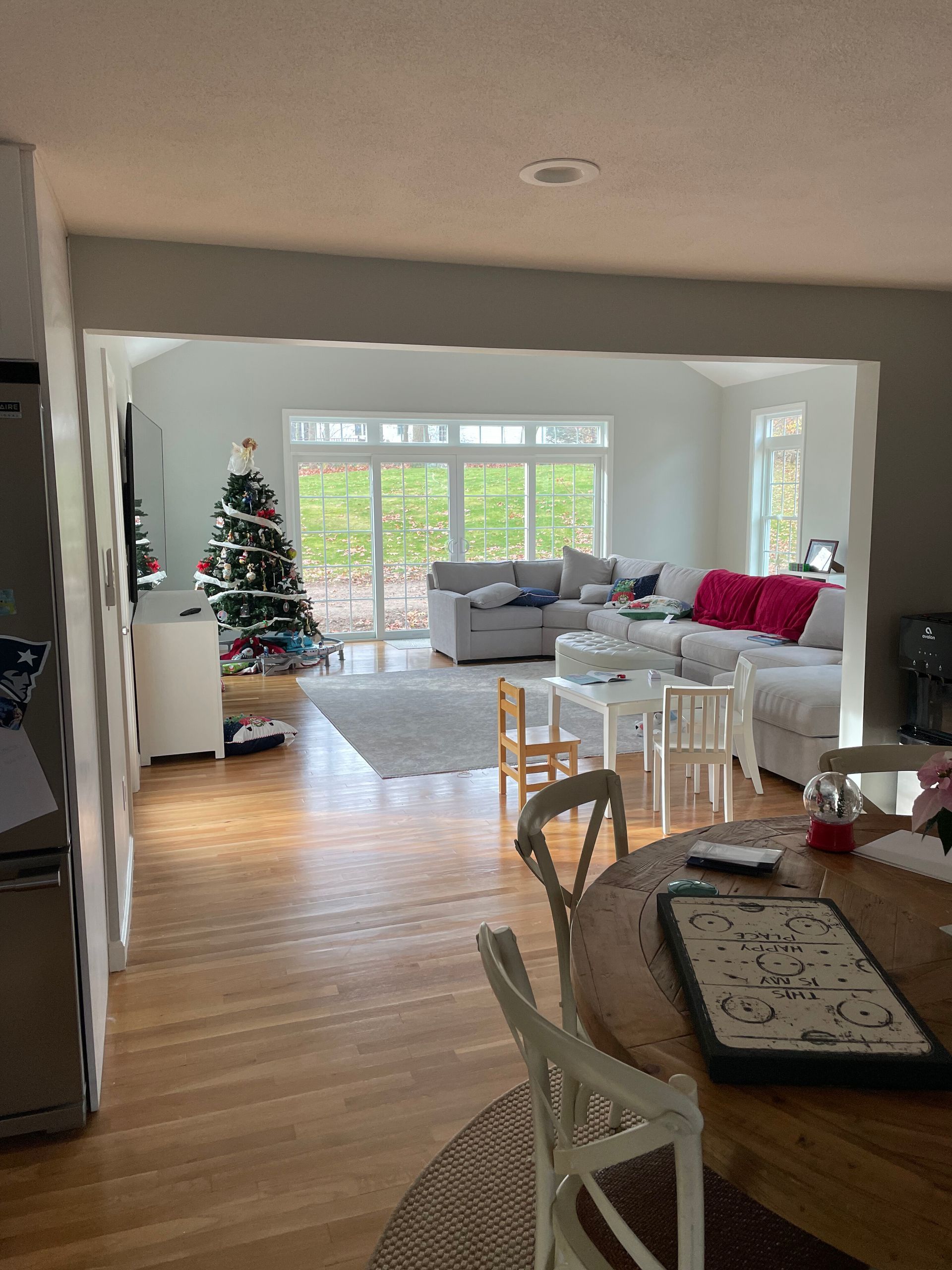 A sunlit open-plan living room with wood floors, a sectional sofa, a Christmas tree, and a round dining table in the front.