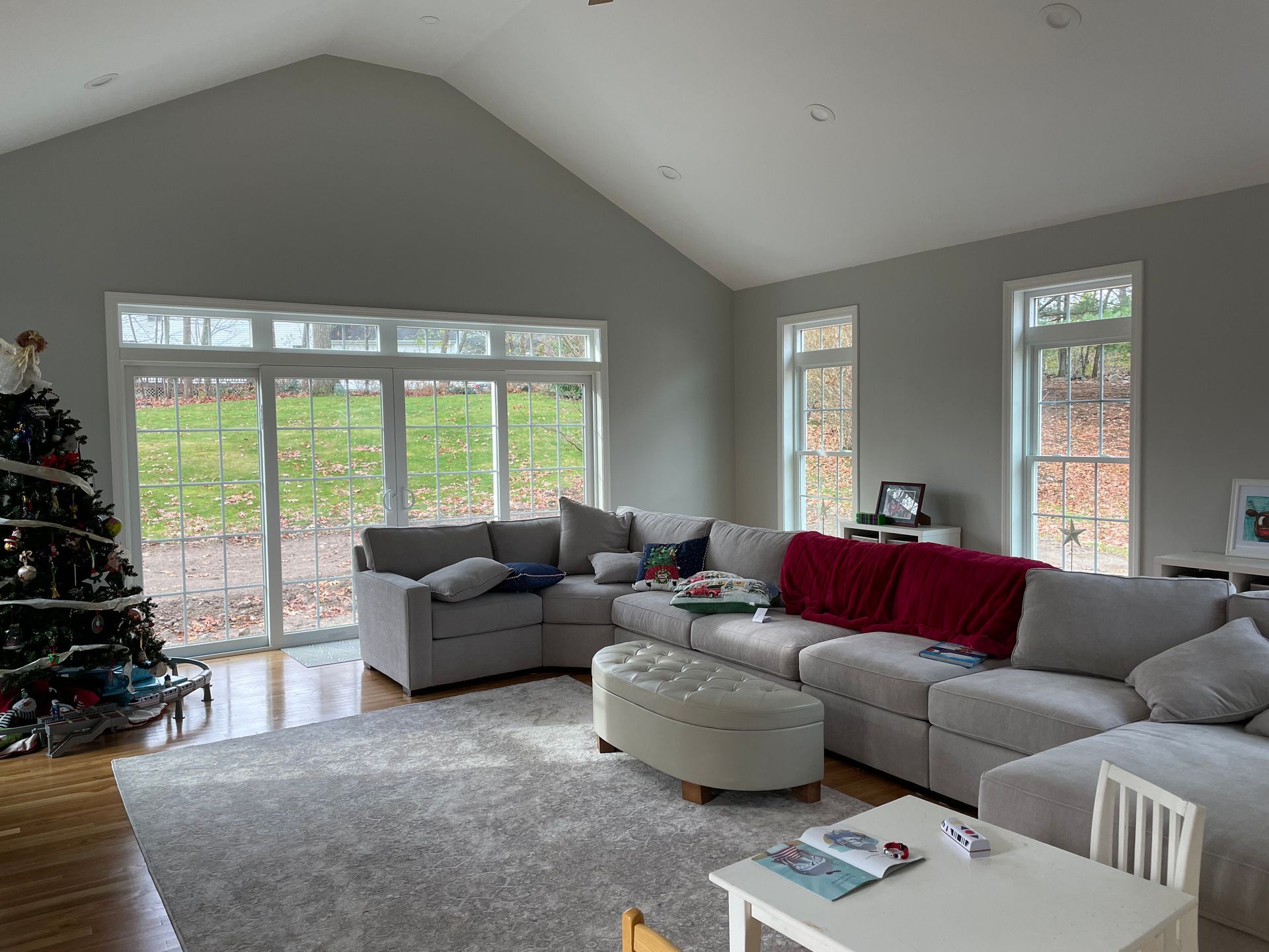 A spacious, bright living room features a large gray sectional sofa, a matching ottoman, and a Christmas tree by windows.