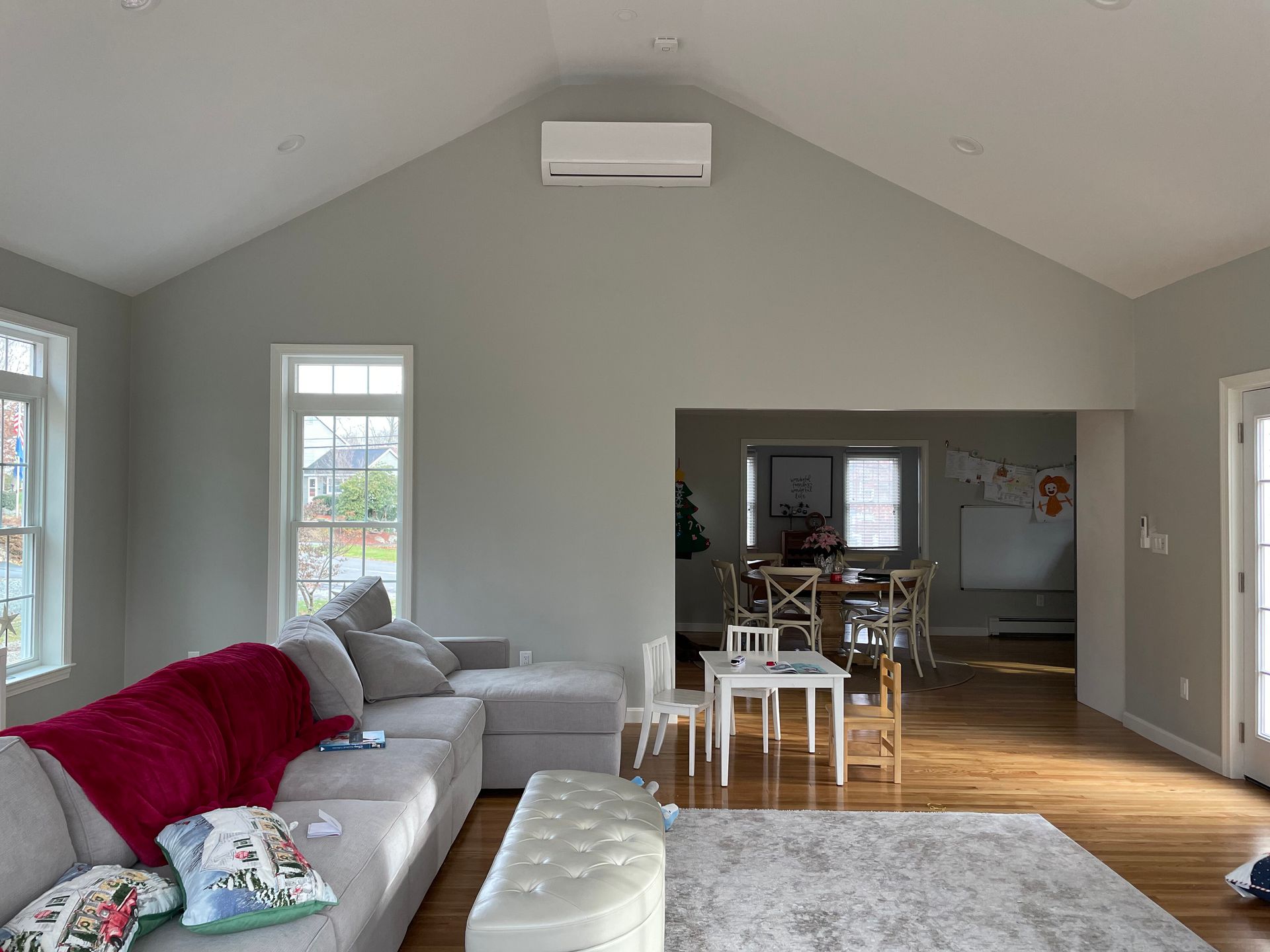 A living room with a large gray sectional, a white ottoman, and a dining area visible through a large wall opening.