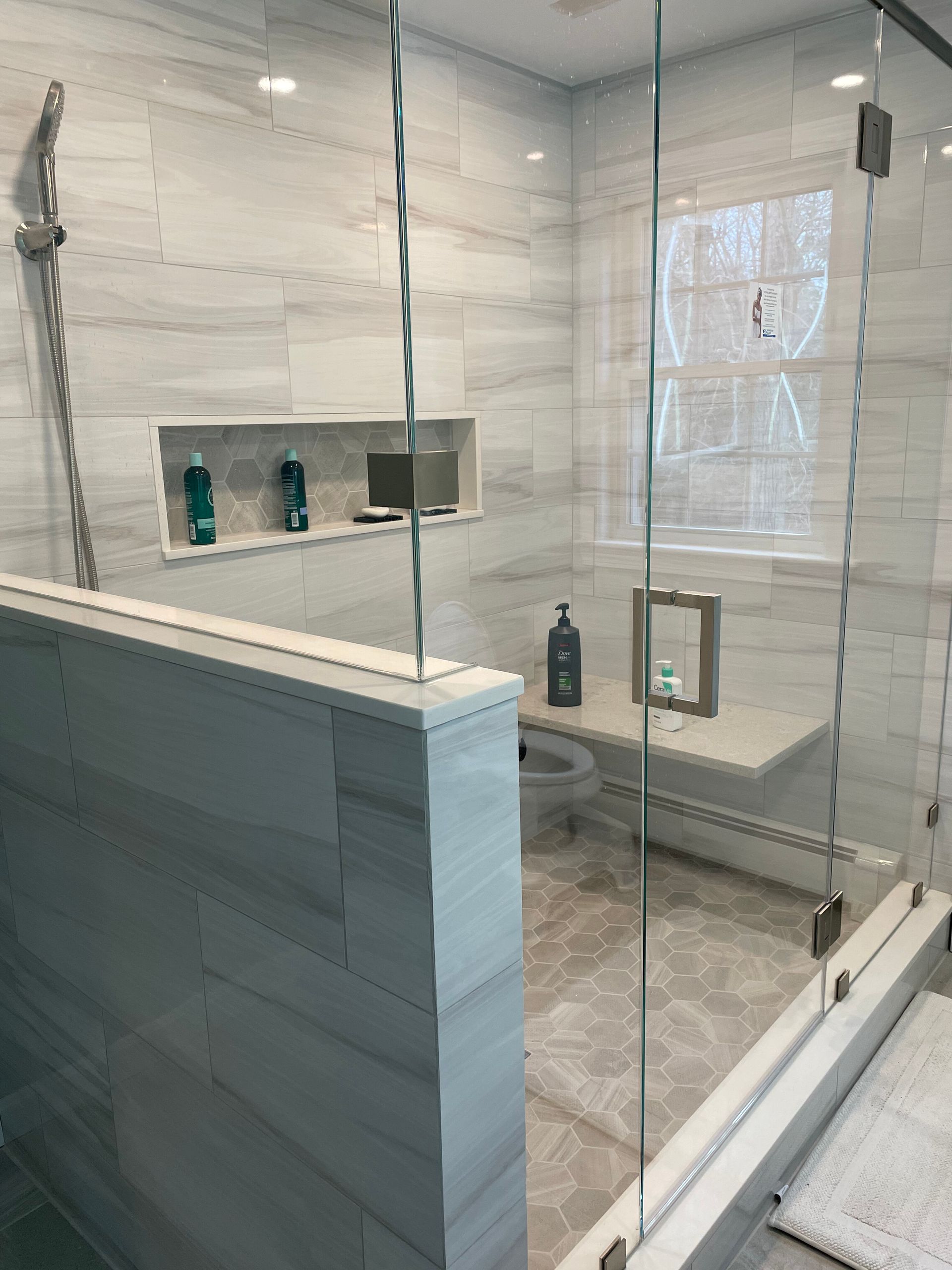 A modern walk-in shower with light grey tile walls, a built-in recessed shelf, a glass enclosure, and a stone bench.