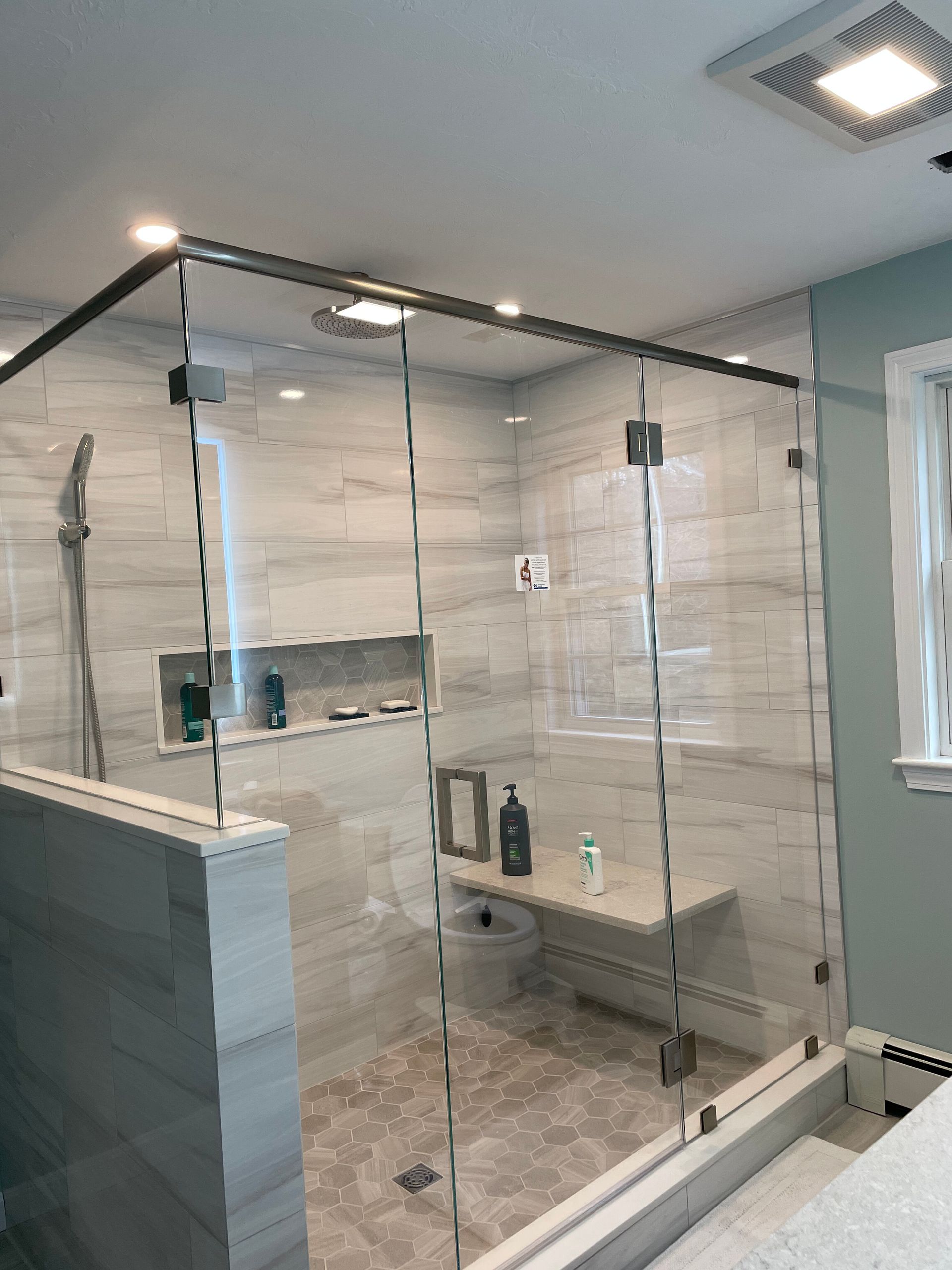 A modern walk-in shower with light gray marble-look wall tiles, a built-in bench, a wall niche, and glass panels.