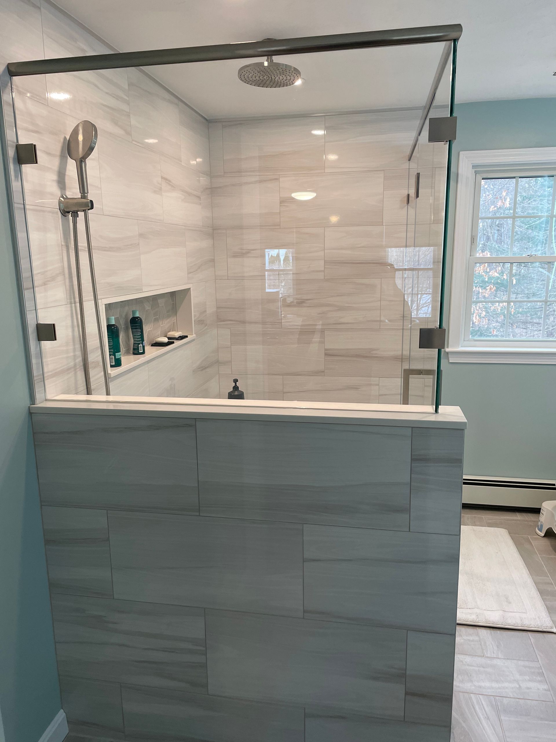 A walk-in shower with light grey stone-look tile, glass panels, a rainfall showerhead, and a handheld wand.