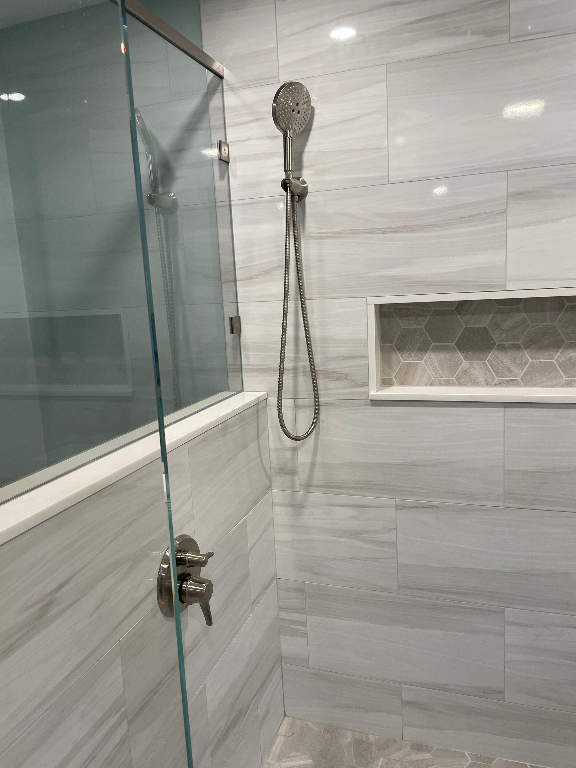 A modern walk-in shower with light gray marble-look tiles, a glass wall, a chrome handheld sprayer, and a tiled niche.