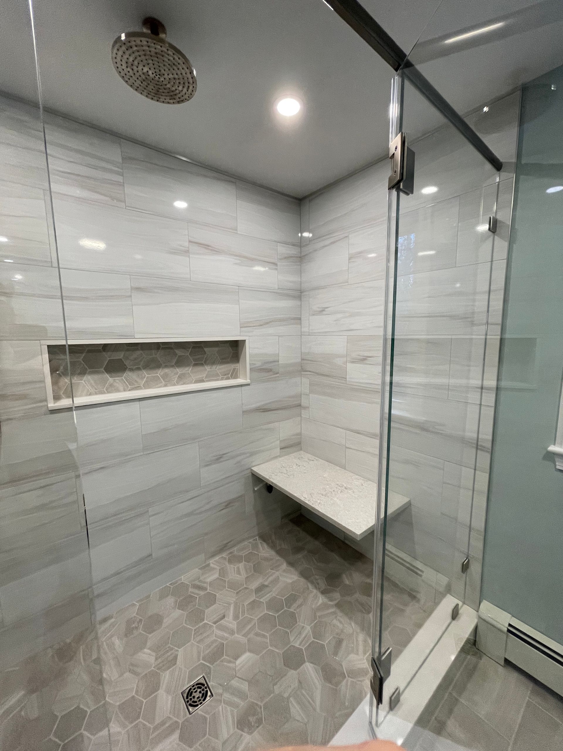 A modern walk-in shower with light grey marble-style tiles, a built-in stone bench, a recessed niche, and a glass door.
