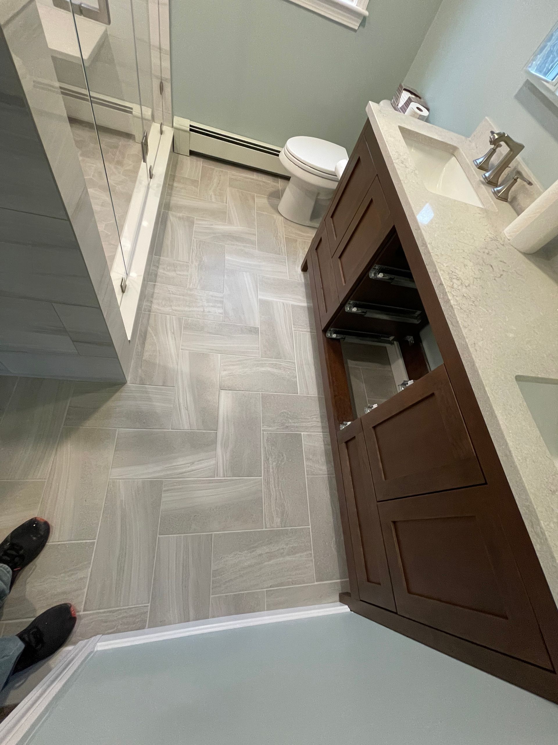 A bathroom renovation with light wood-grain tile in a herringbone pattern, a dark wood vanity, and a glass-enclosed shower.