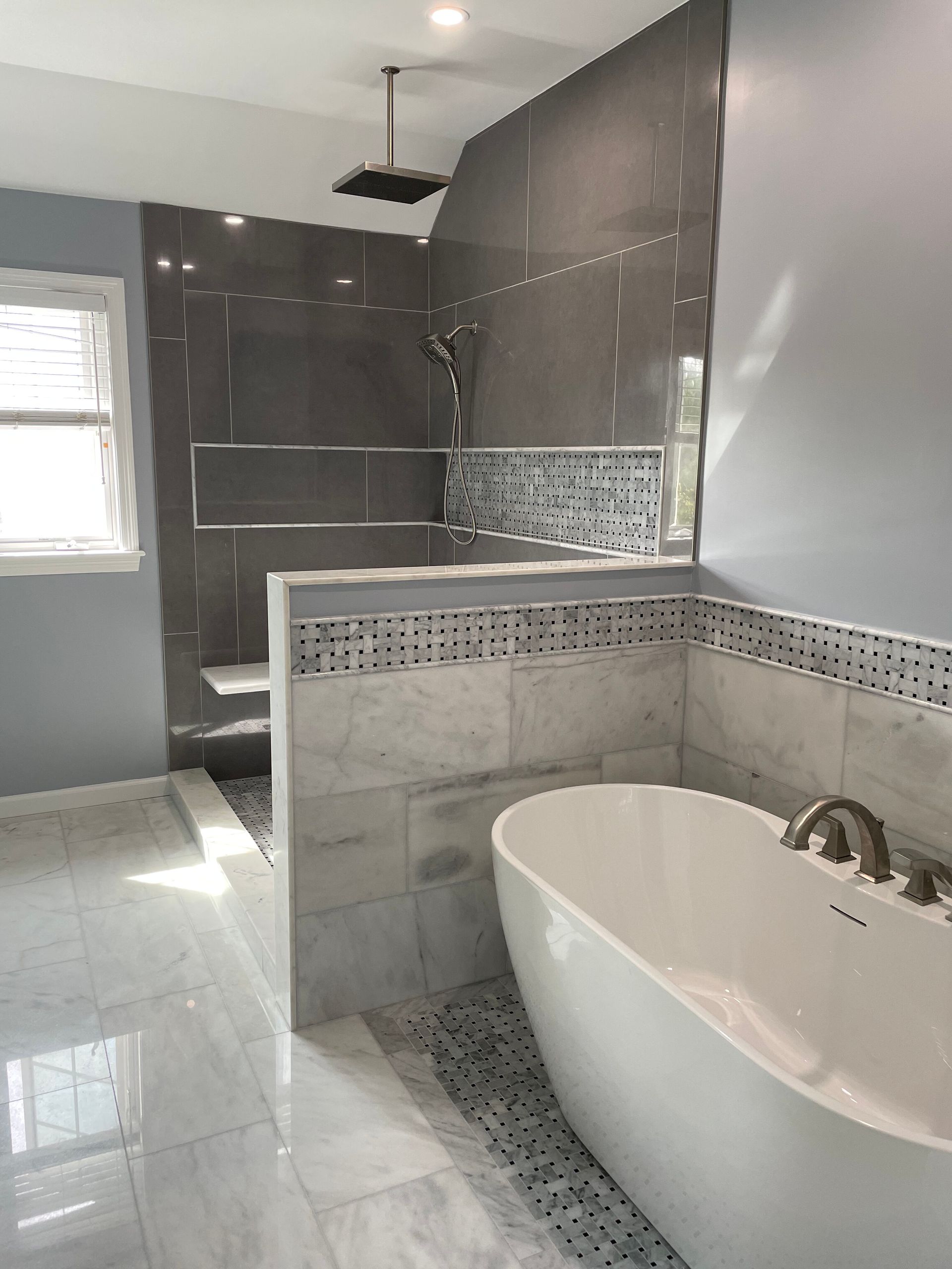 A modern bathroom featuring a white freestanding tub, a marble-tiled half wall, and an open glass-enclosed shower area.