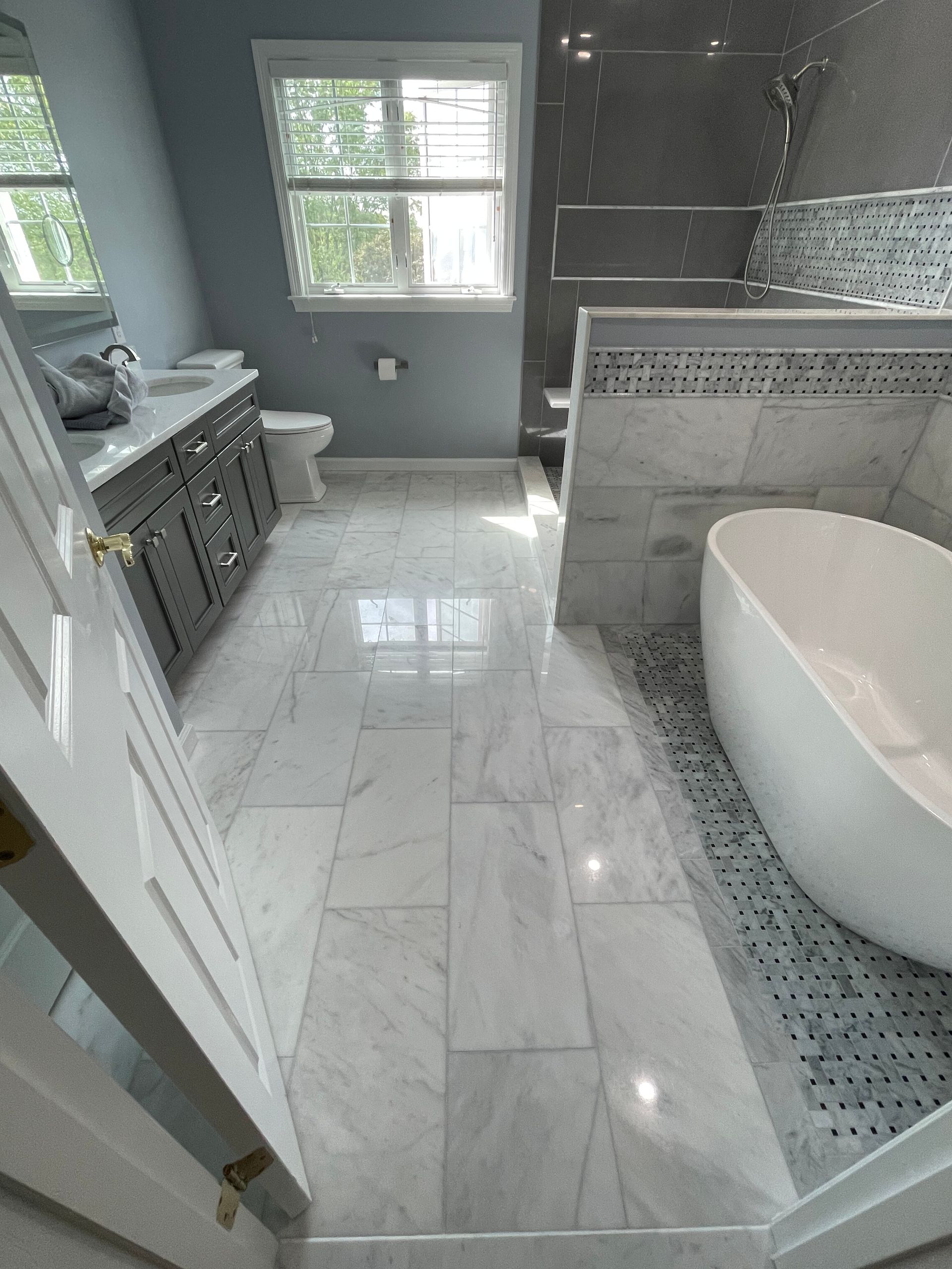 A renovated bathroom with a grey vanity, a white soaking tub, light-colored marble flooring, and a glass-enclosed shower.