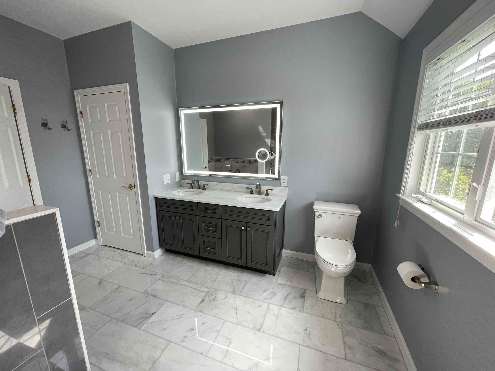 A modern bathroom with grey walls, white marble floors, a dark vanity, a lit vanity mirror, and a white toilet.