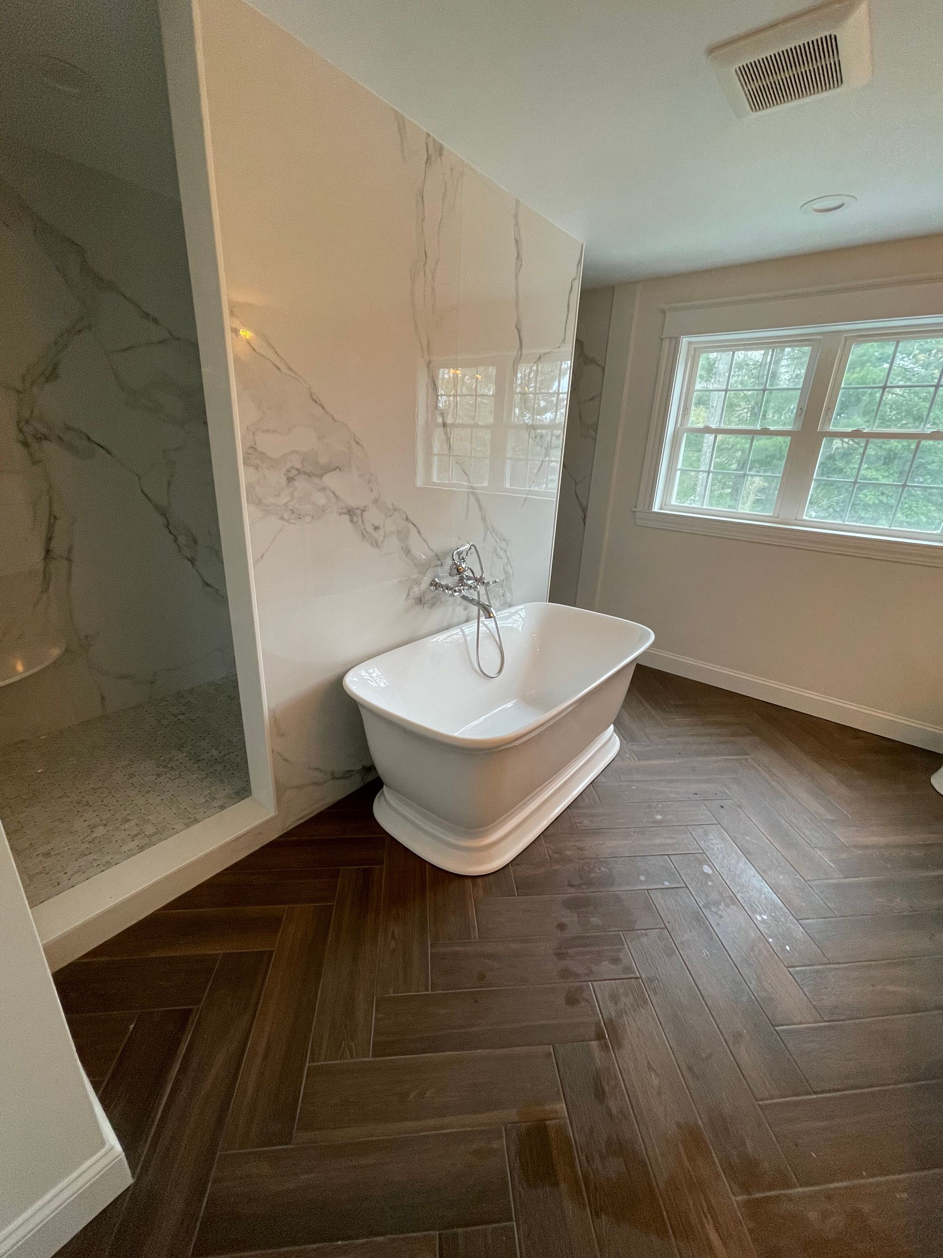 A modern bathroom featuring a white freestanding tub against a large marble slab wall, next to a shower and wood flooring.