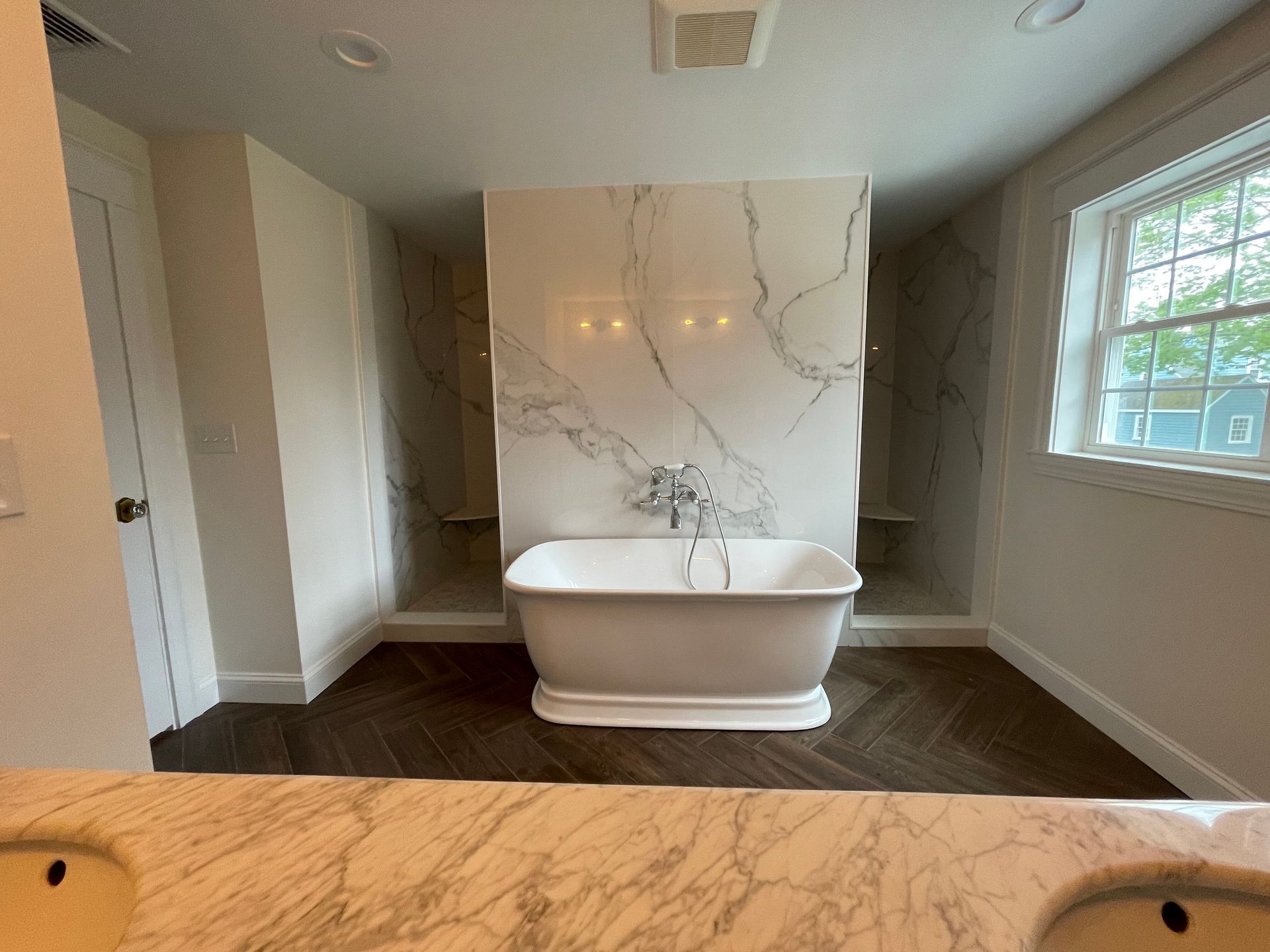 A freestanding bathtub centered in a bathroom with marble-tiled walls and a herringbone-patterned floor.