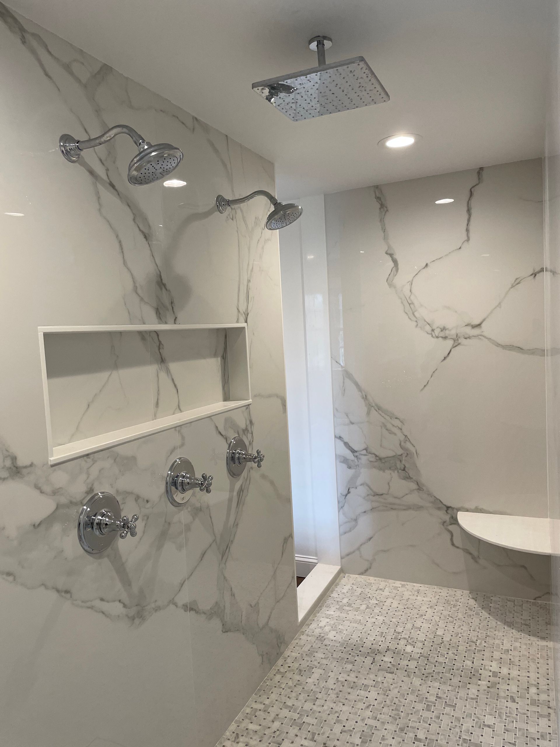 A modern shower with white, grey-veined marble walls, a built-in shelf, two shower heads, a rain shower, and a corner seat.