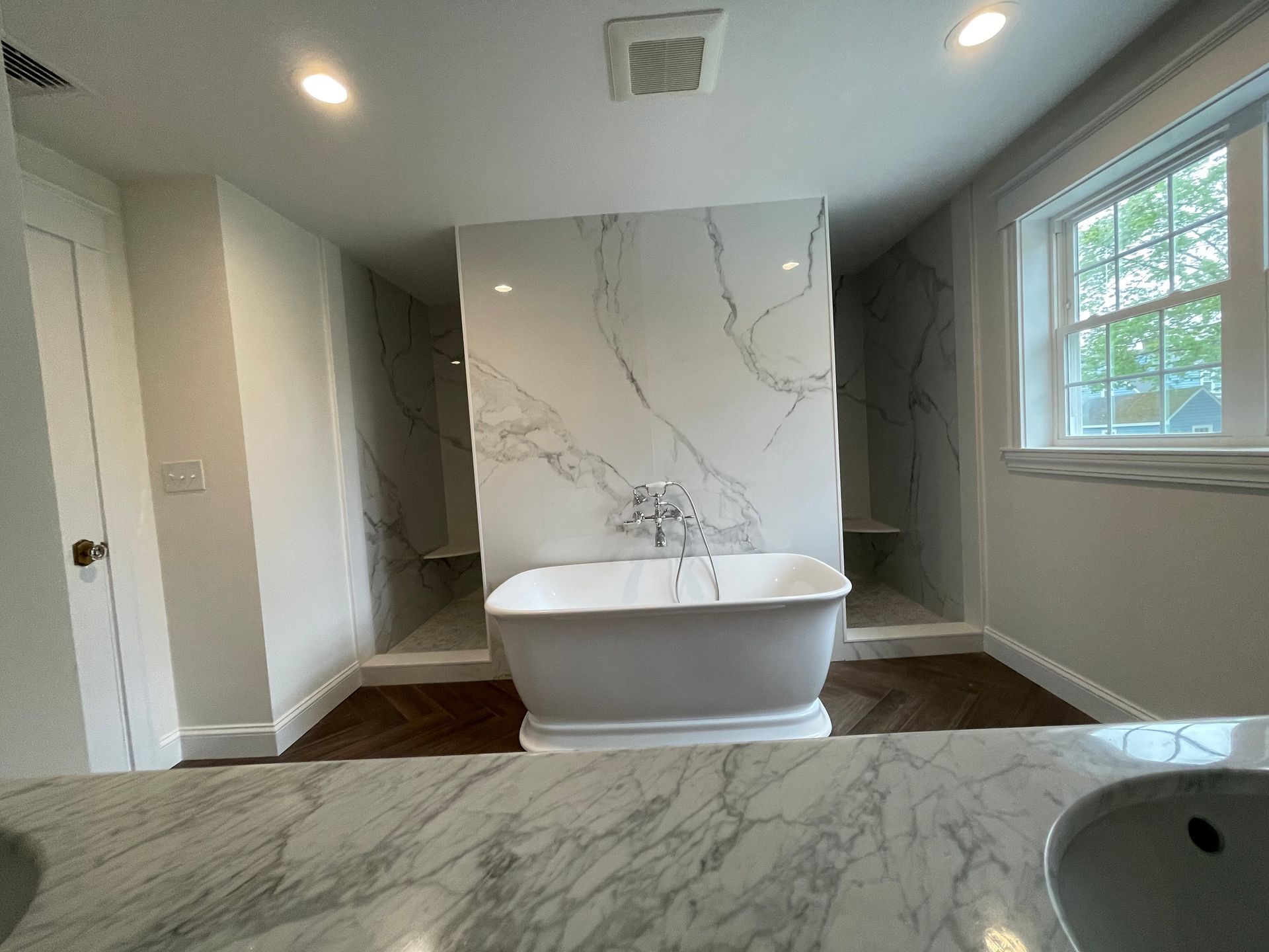 A modern bathroom featuring a white freestanding tub in front of a marble-tiled wall, flanked by two walk-in showers.