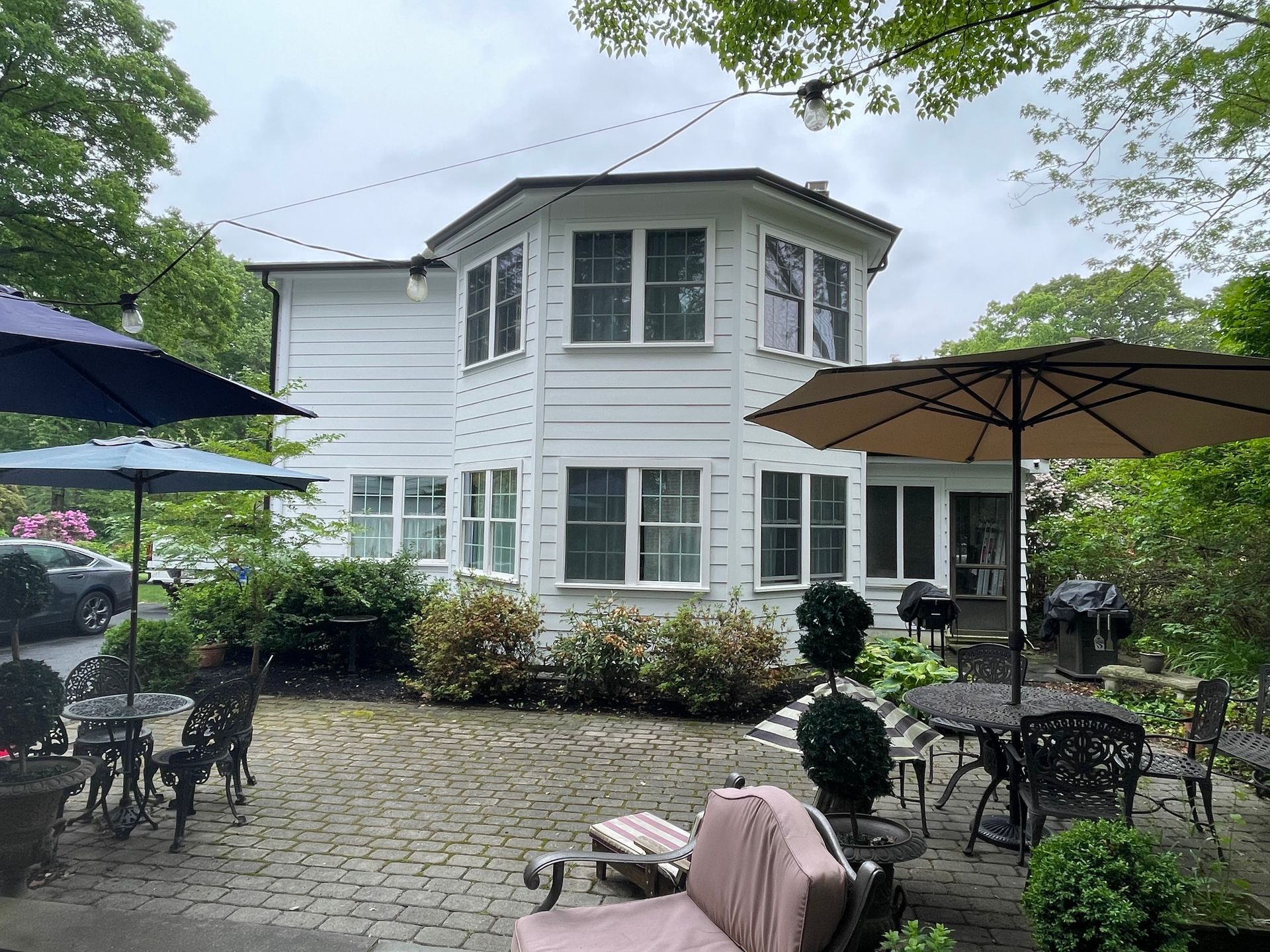 A white, two-story house with a rounded exterior view stands behind a cobblestone patio featuring outdoor café seating.