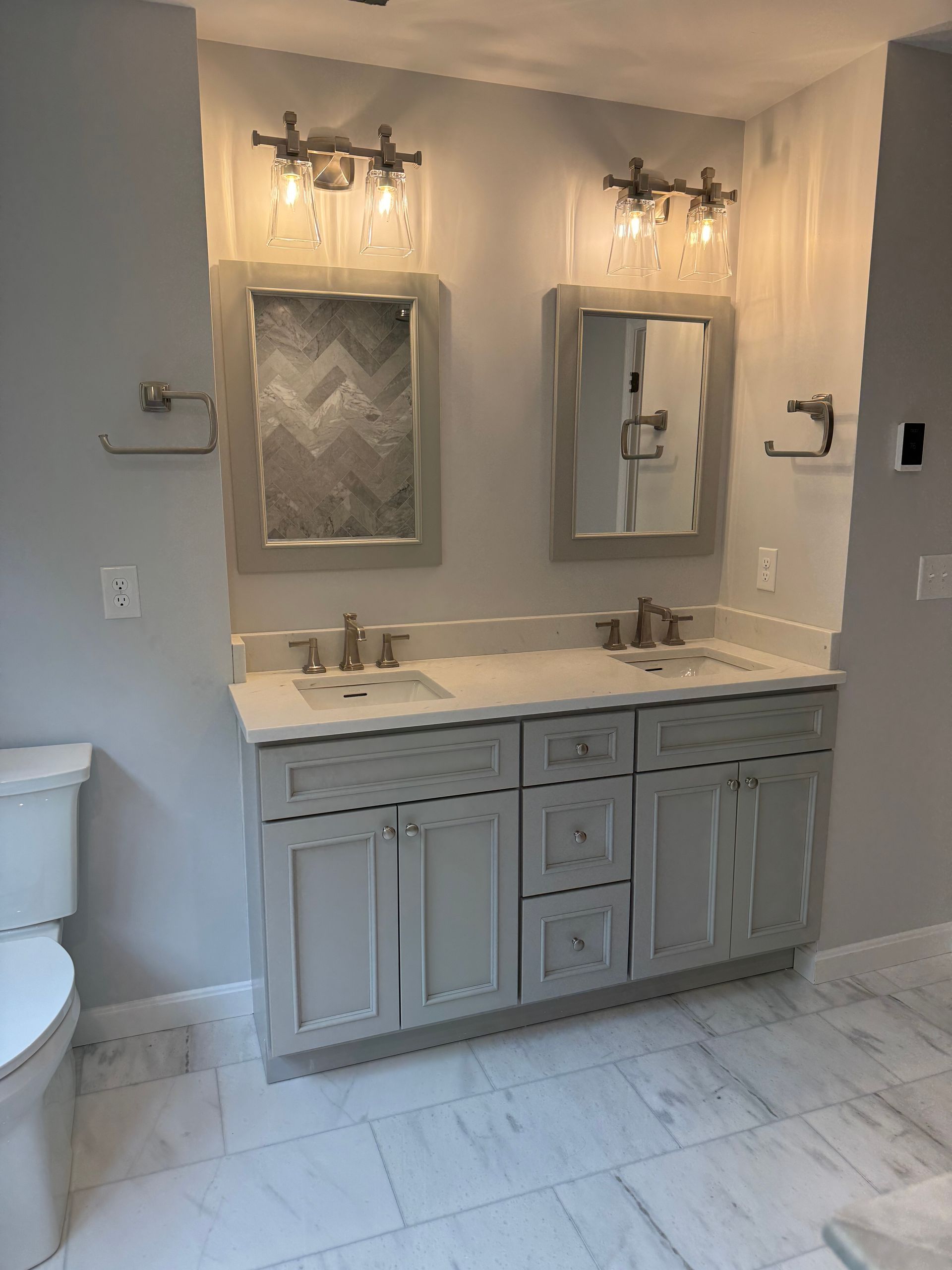 A bathroom vanity with two sinks, gray cabinets, marble-patterned floors, two mirrors with sconces, and light gray walls.