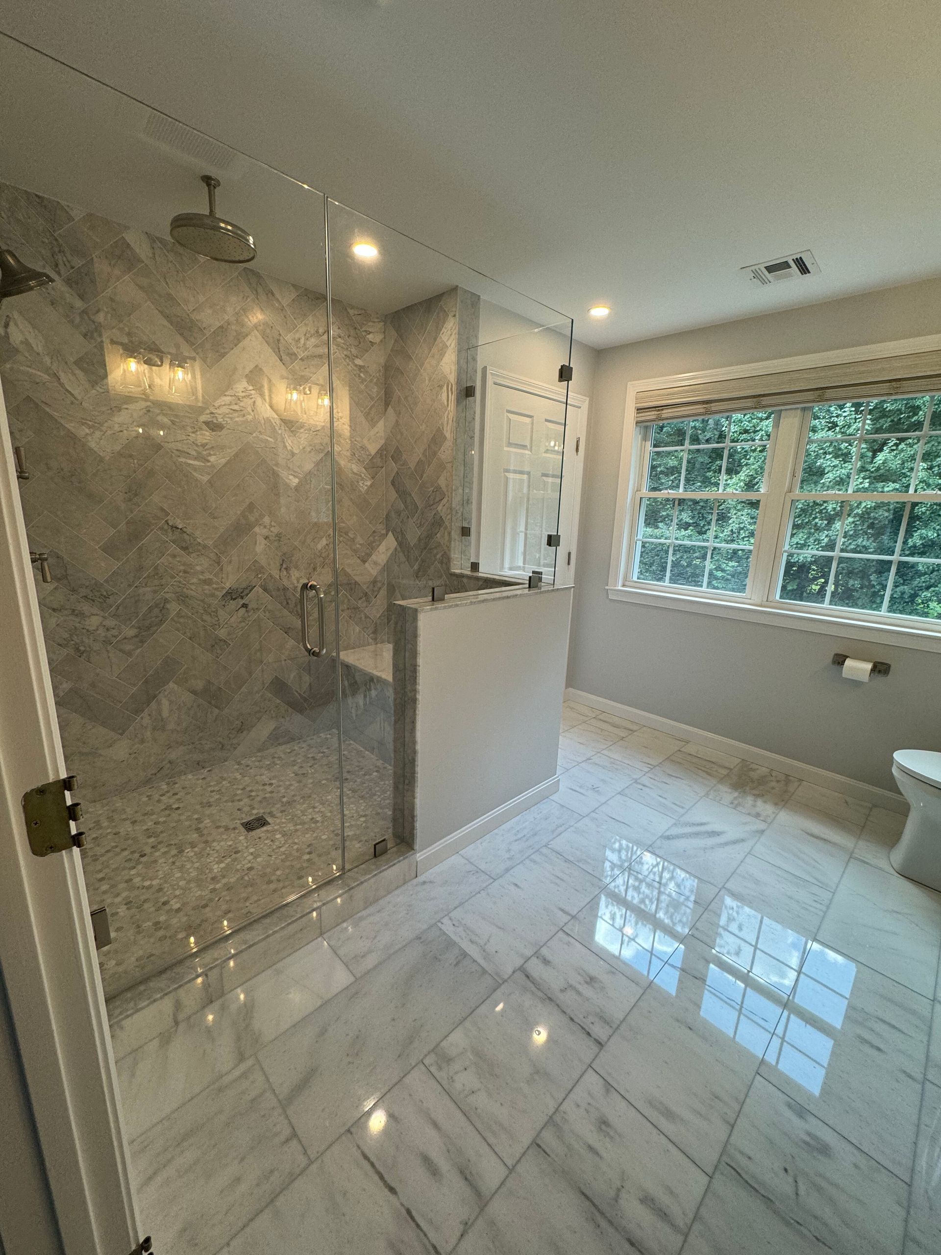 A bright bathroom with marble tile floors, a glass-enclosed shower with chevron walls, and a window with white shutters.