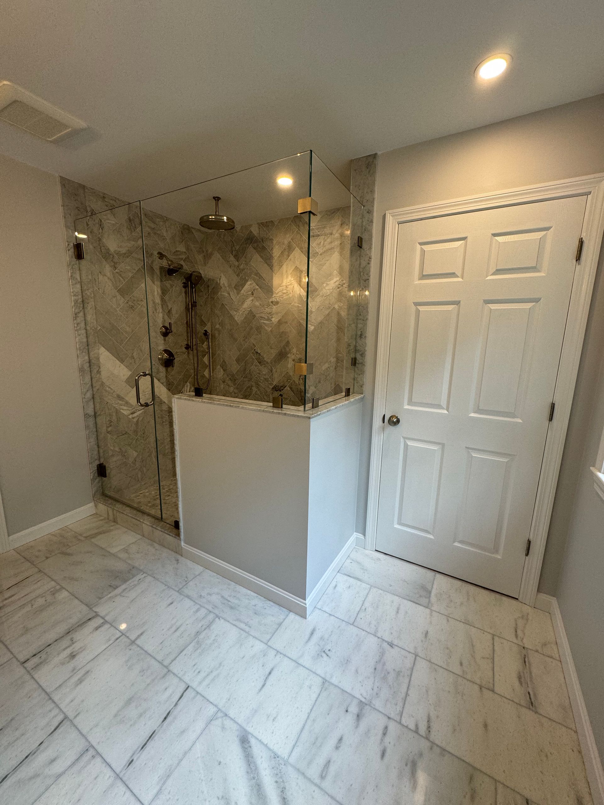 A bathroom with white marble floors, a glass-enclosed shower with grey patterned tile, and a white six-panel door.