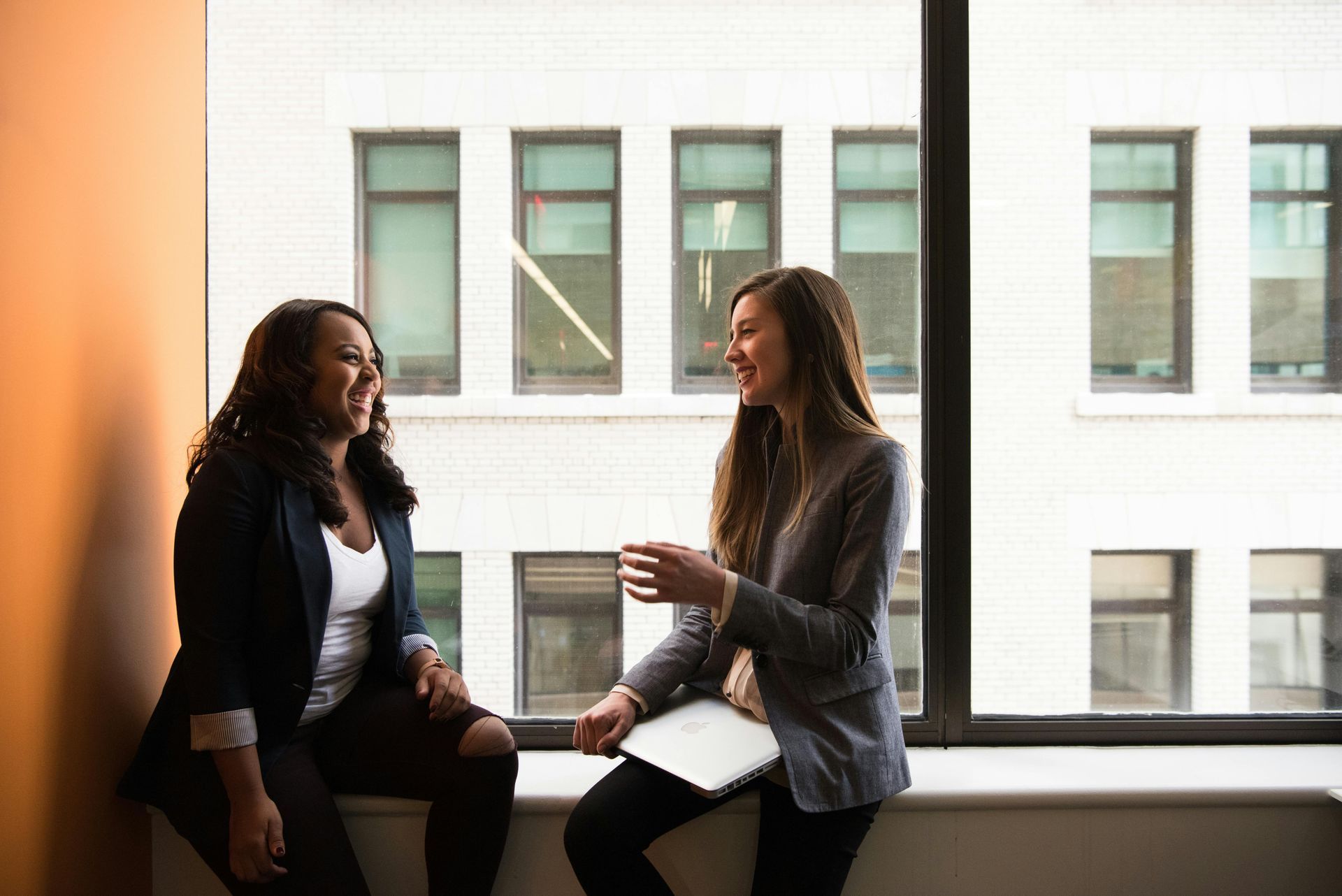 Two women are sitting on a window sill having a conversation.
