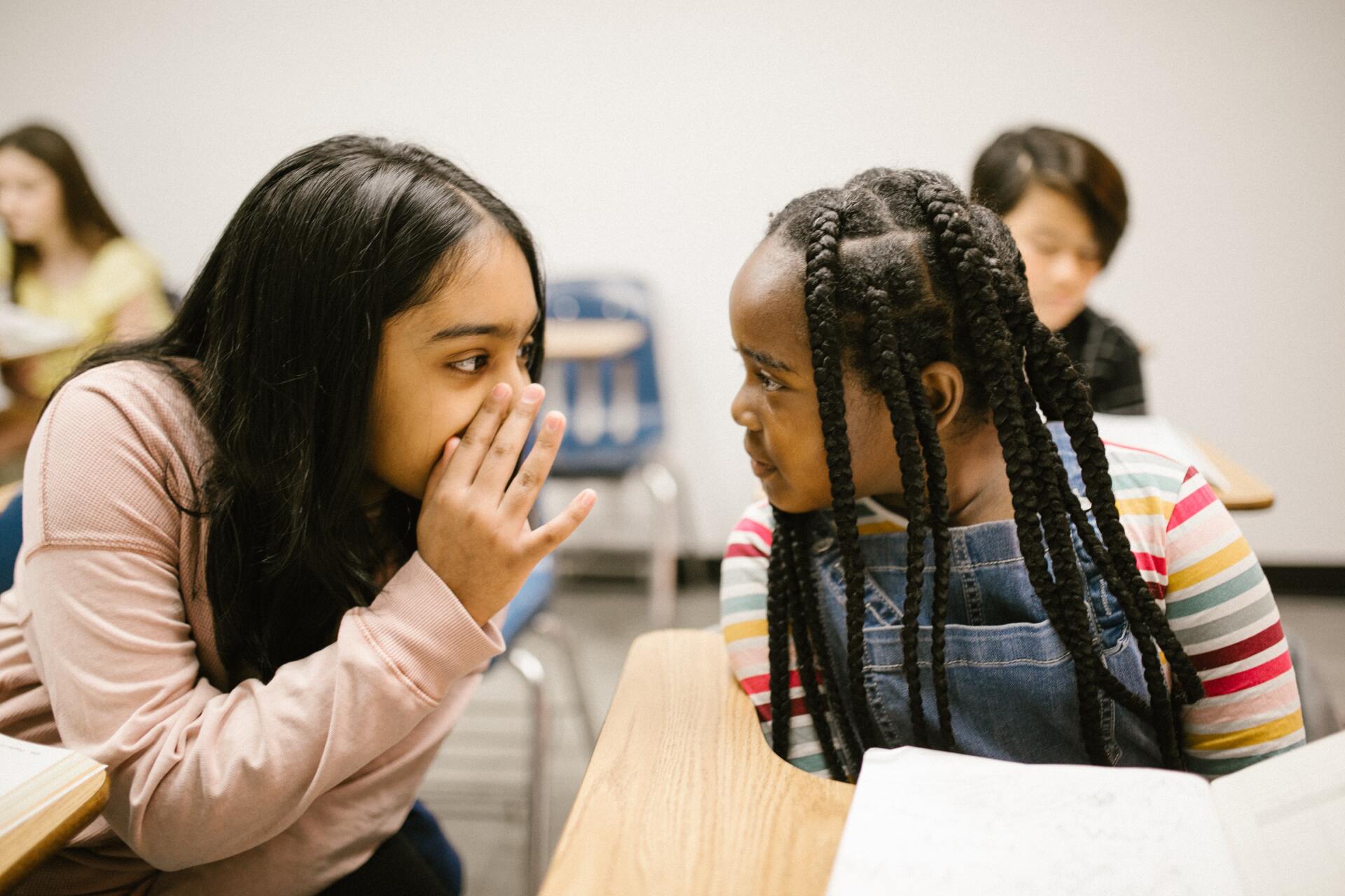 A student with black hair whispers to another student with braids