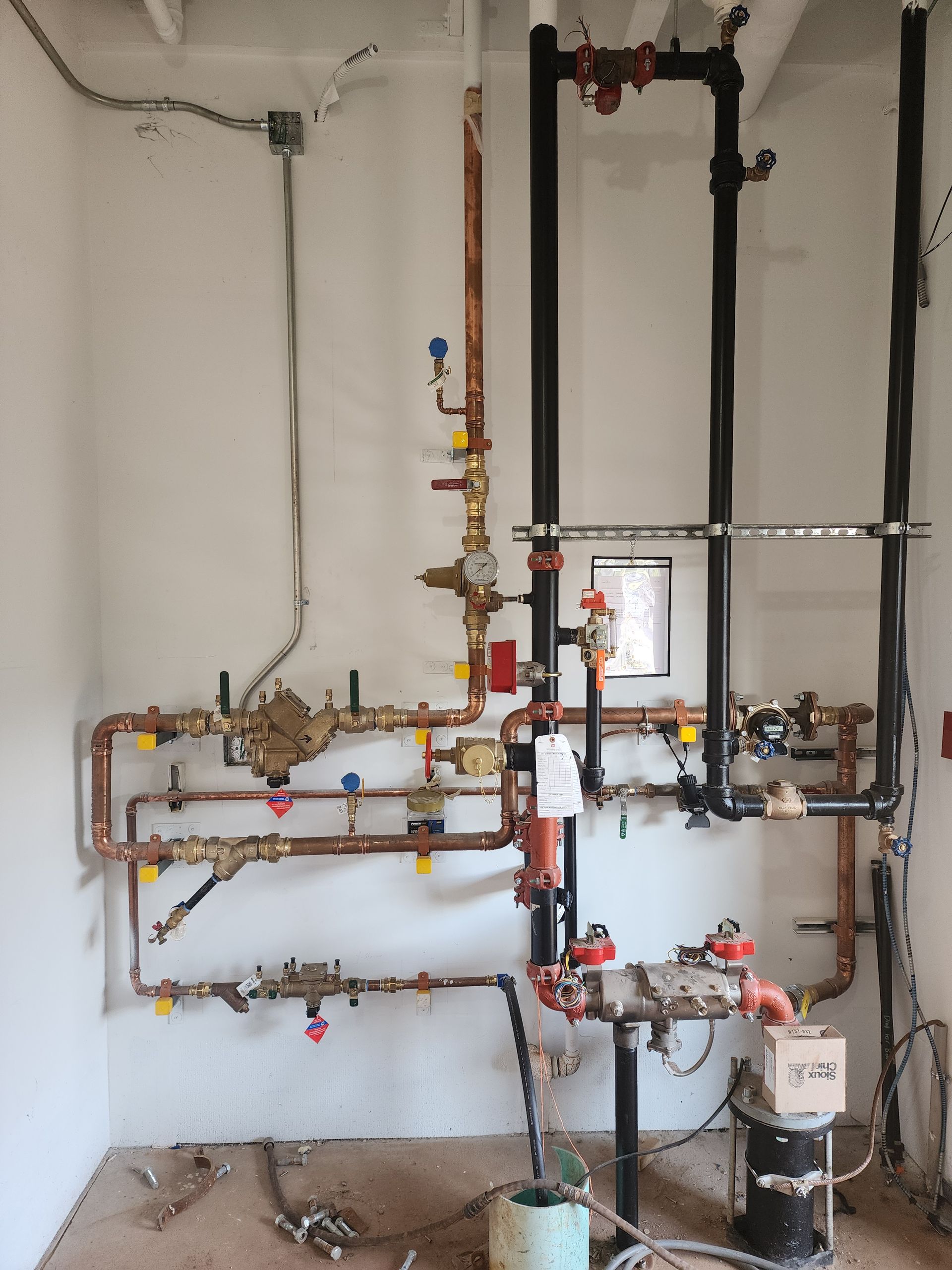 Wall-mounted plumbing and heating pipes with valves in a utility room, mostly metal with copper and black lines.