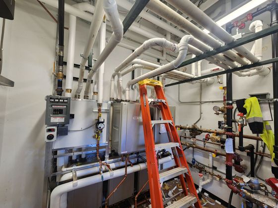 Industrial room with white pipes, an orange ladder, and a worker in a yellow safety vest adjusting equipment.