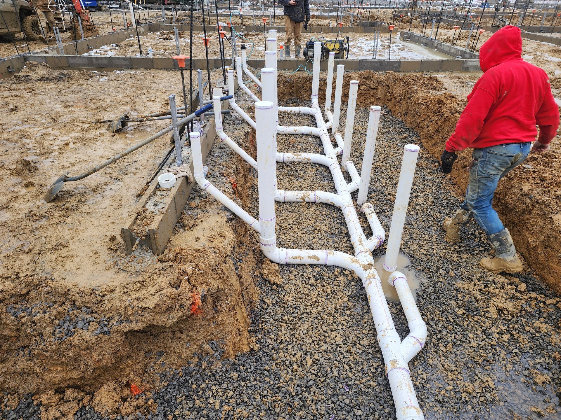 Construction site with white pipes being installed in a trench, workers in the background and a person in a red hoodie nearby