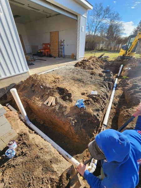 Person in blue hoodie digging a trench beside a garage with a shovel.