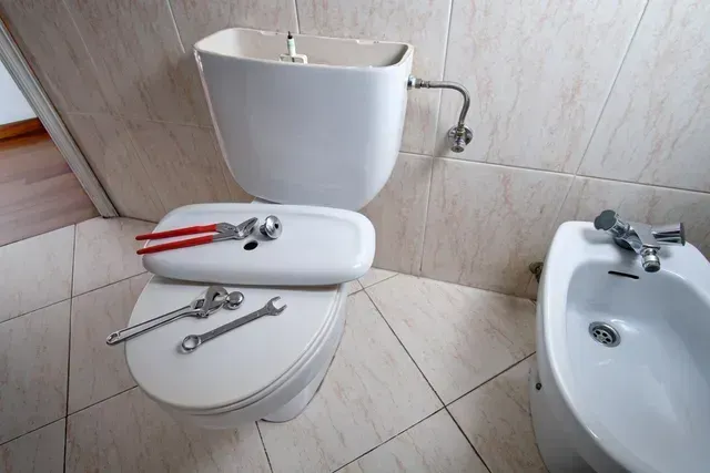 Bathroom with a toilet seat covered in pliers and wrenches next to a bidet and toilet tank