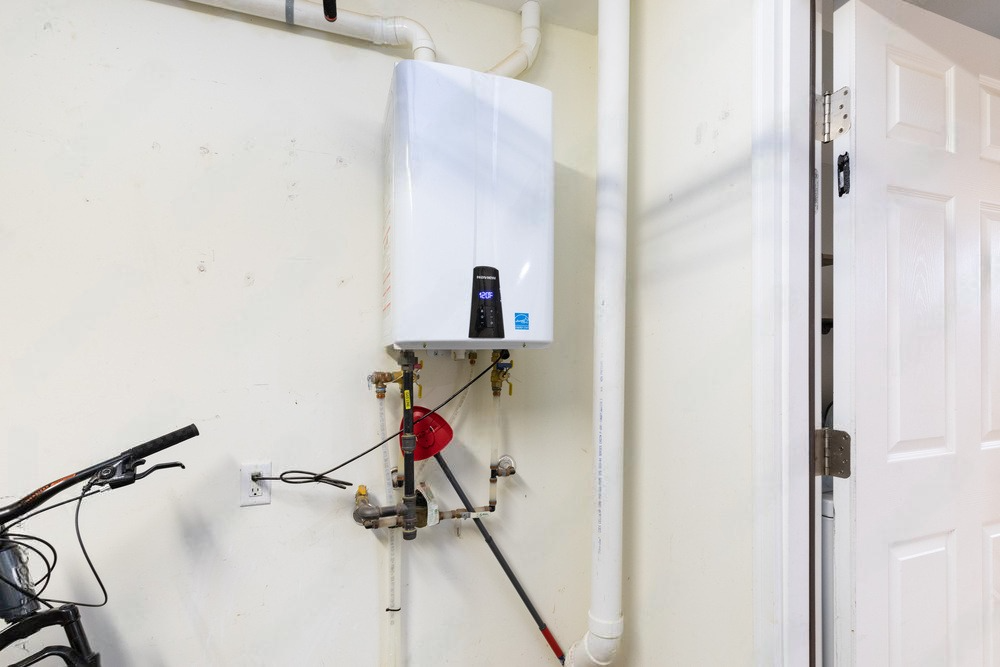 White water heater in garage beside pipes, valve, and a door, with a bike handlebar at left.
