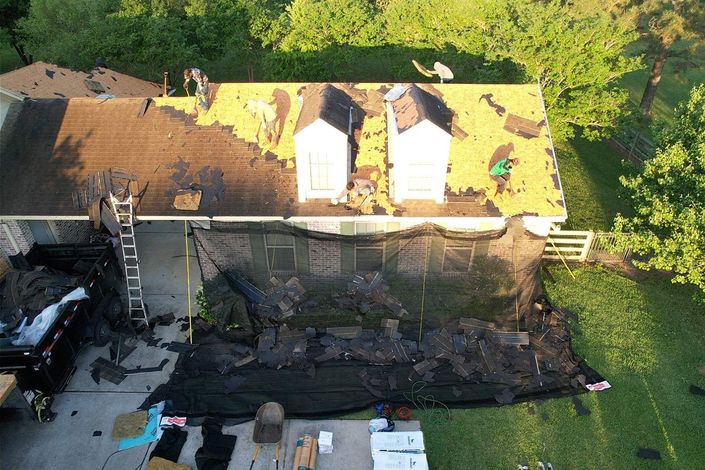 Roofer in hard hat and safety vest inspecting shingles, taking notes on clipboard.