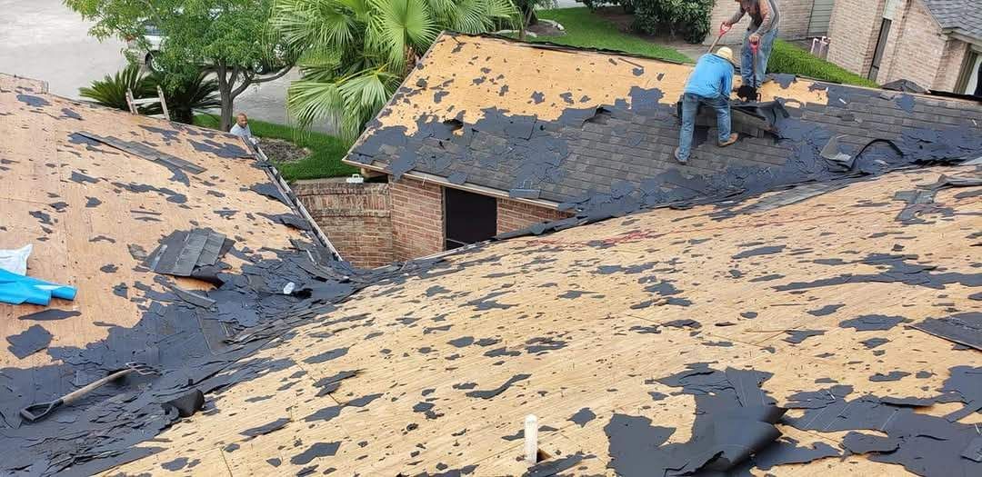 Roofer inspecting a shingled roof with a tool; he kneels, wearing a safety vest and cap.