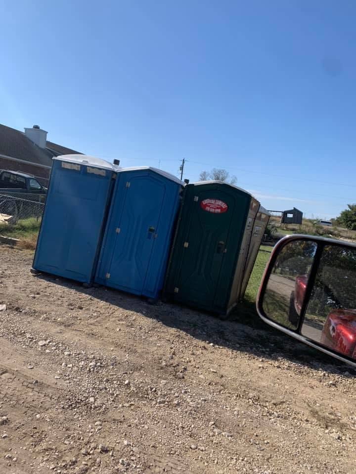 Three portable toilets, two blue and one dark green, on a gravel lot under a clear blue sky.