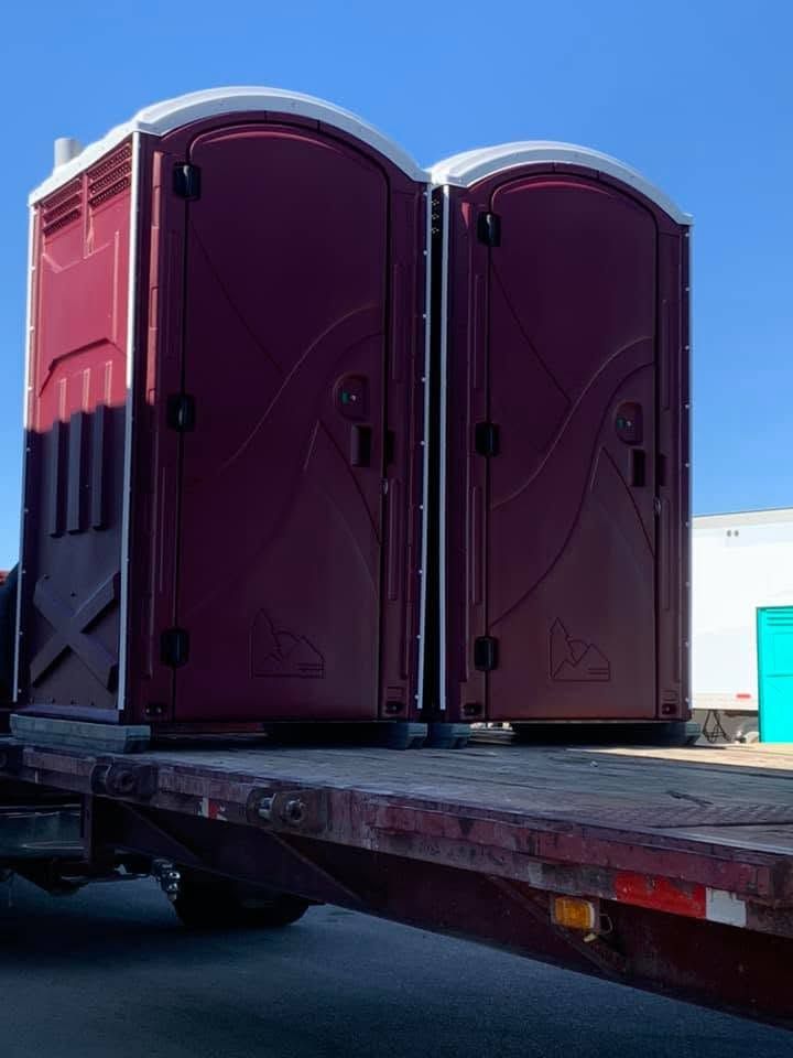 Two maroon portable toilets on a flatbed trailer under a blue sky